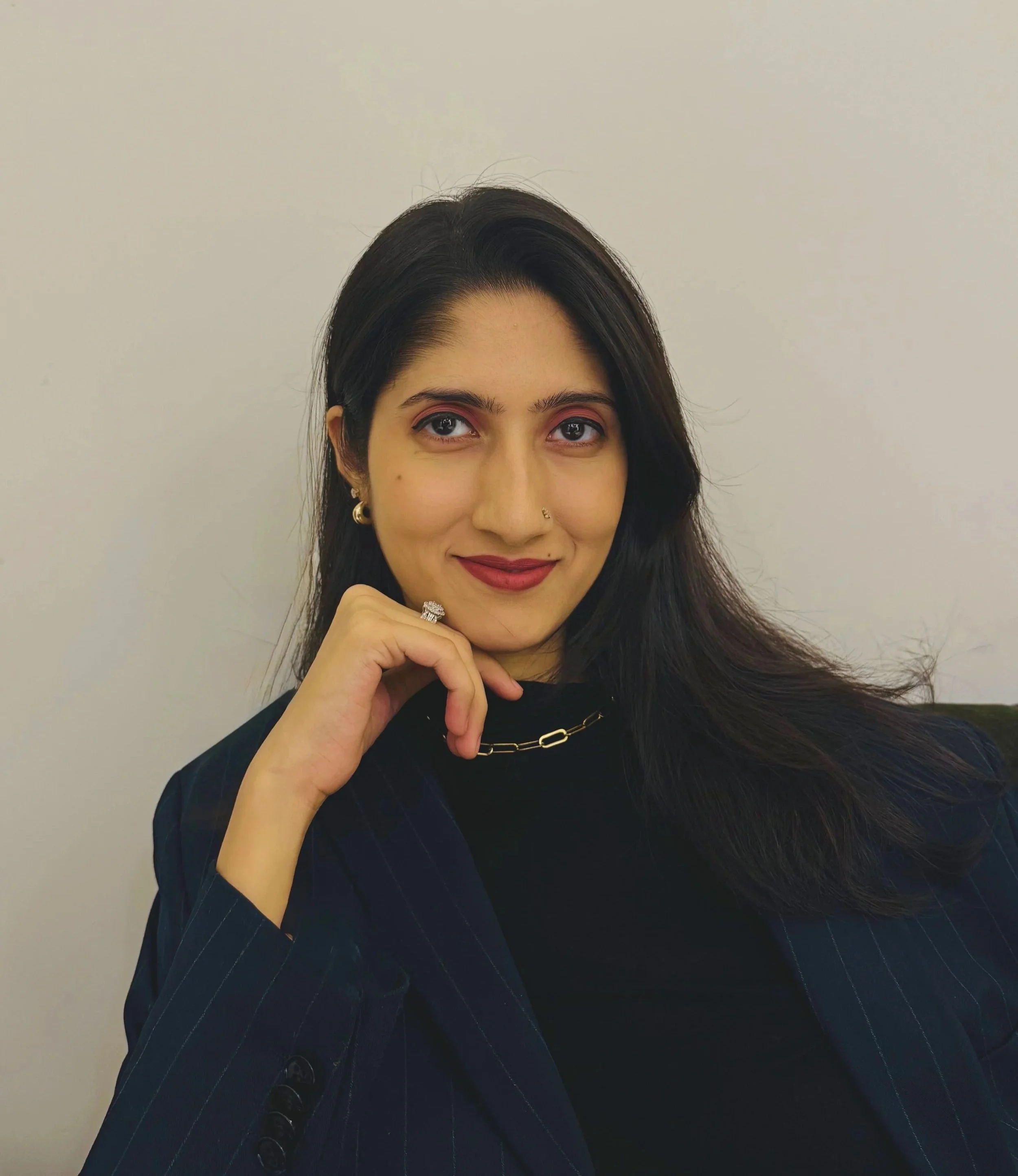 A woman with long black hair, wearing a black top and a blue pinstripe blazer, smiling with her chin resting on her hand, standing against a plain white background.
