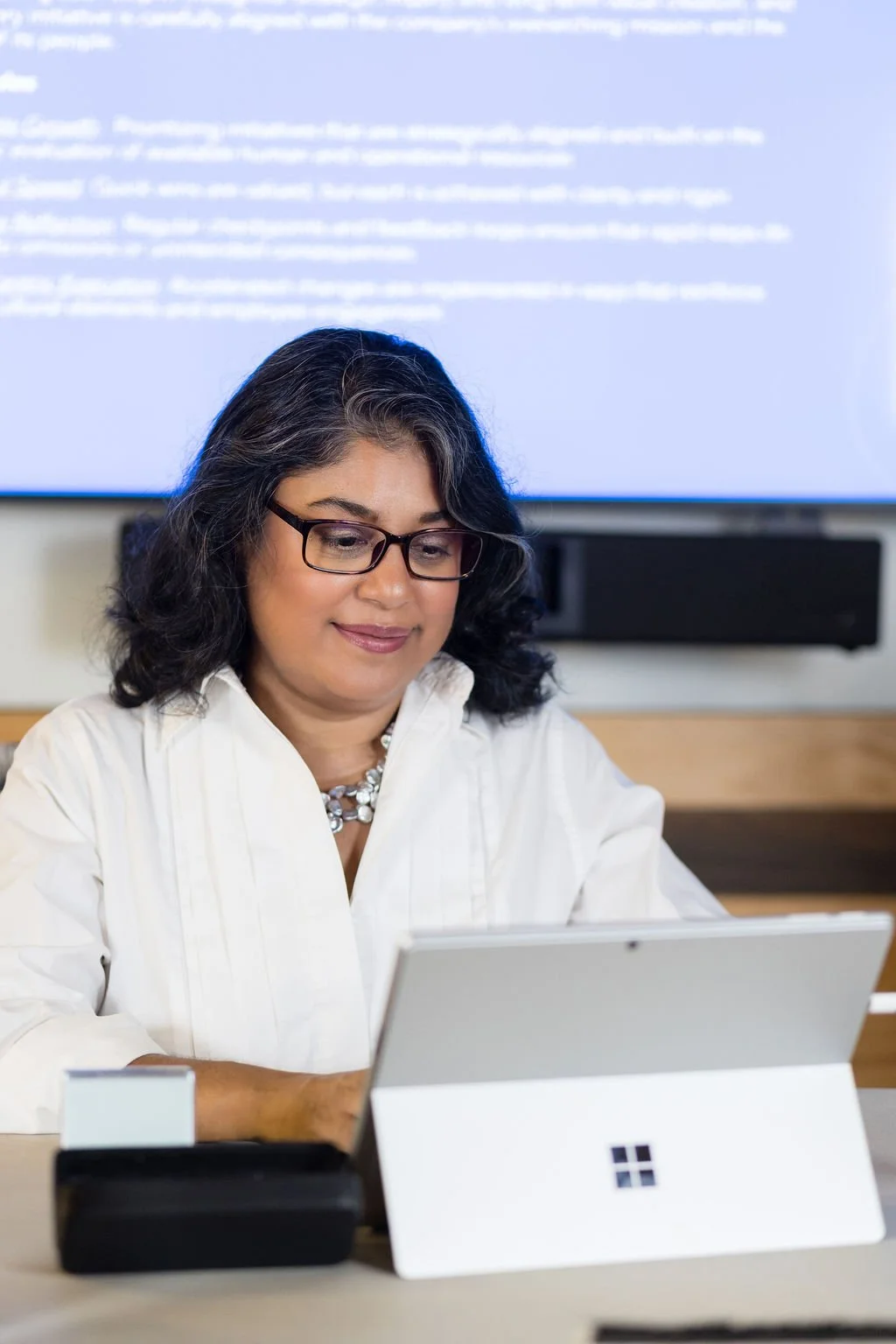 A woman with glasses and dark hair, wearing a white blouse and pearl necklace, working on a tablet at a desk with a Microsoft Surface device, in front of a large screen displaying blurred text.
