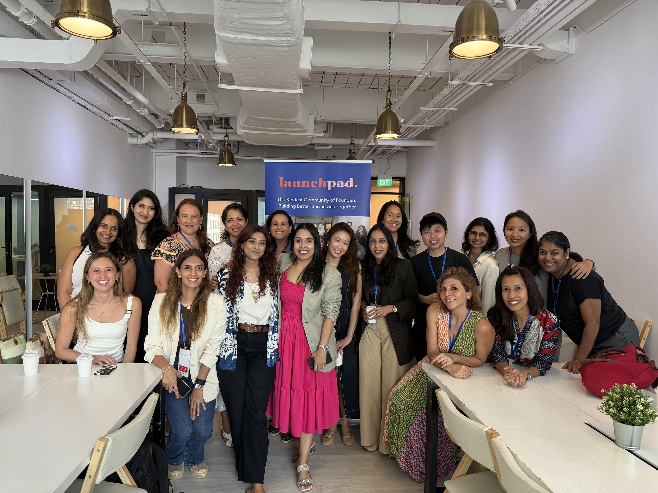 Group of women gathered in a modern office space for a photo, with a banner behind them that reads 'launchpad' and 'The kindest community of founders building better businesses together.' The women are smiling and wearing a mix of casual and business attire.