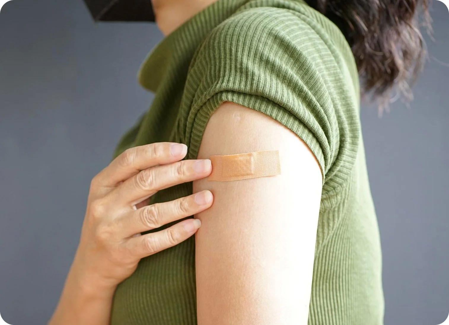 Close-up of a person with dark, wavy hair receiving a vaccination shot in their upper arm, which is covered with a green short-sleeve shirt. A healthcare worker's hand is holding the person's arm, and a bandage is placed over the injection site.