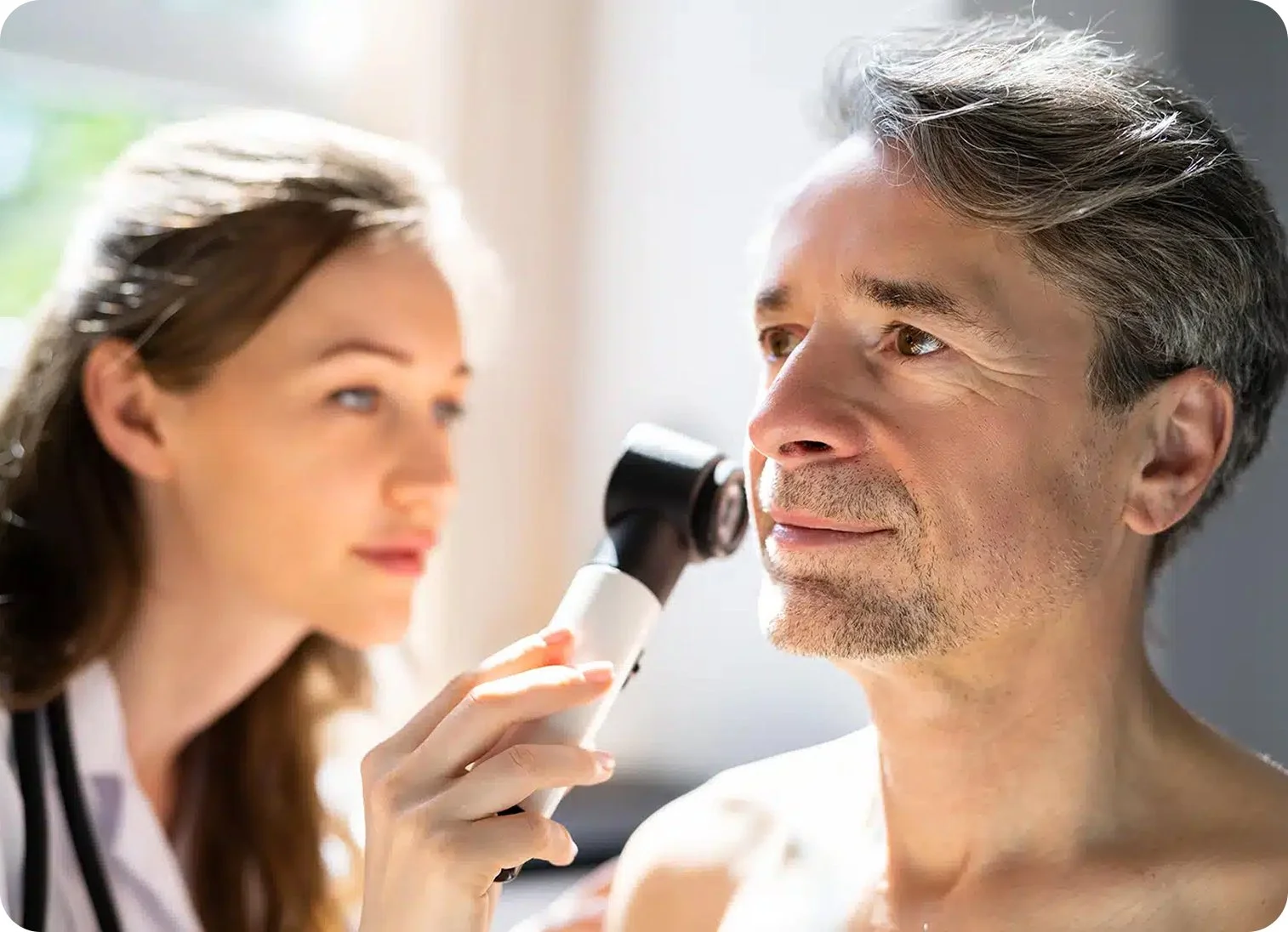 A female doctor examines a middle-aged male patient's ear or throat with an otoscope in a clinical setting.