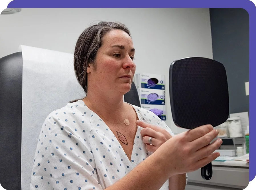 A woman in hospital gown looking at her reflection in a mirror with a tattoo on chest visible, in a medical setting.