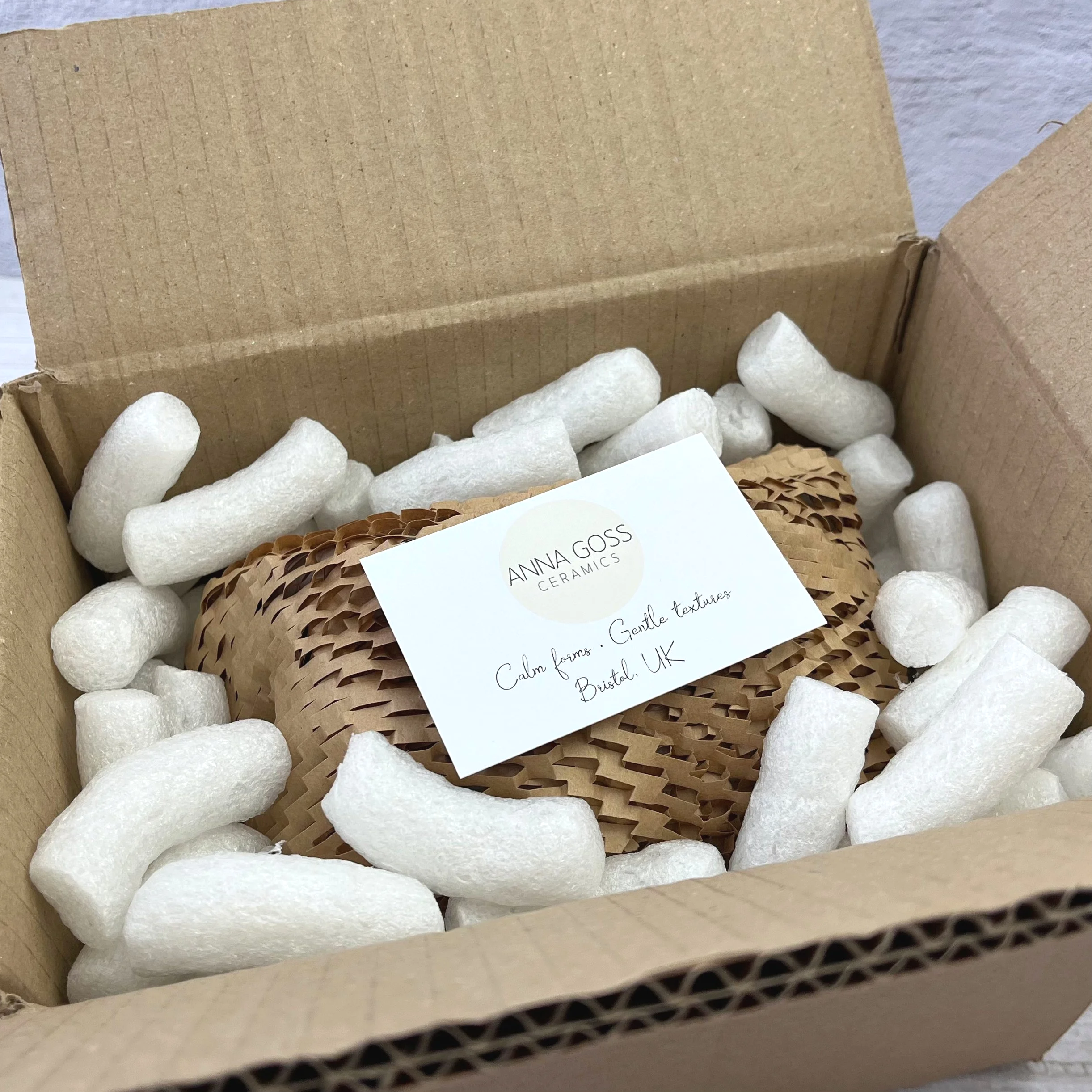 A photo showing how orders are packaged. Open cardboard box filled with white packing peanuts, a decorative ceramic piece wrapped in brown honeycomb paper with a label from Anna Goss Ceramics.