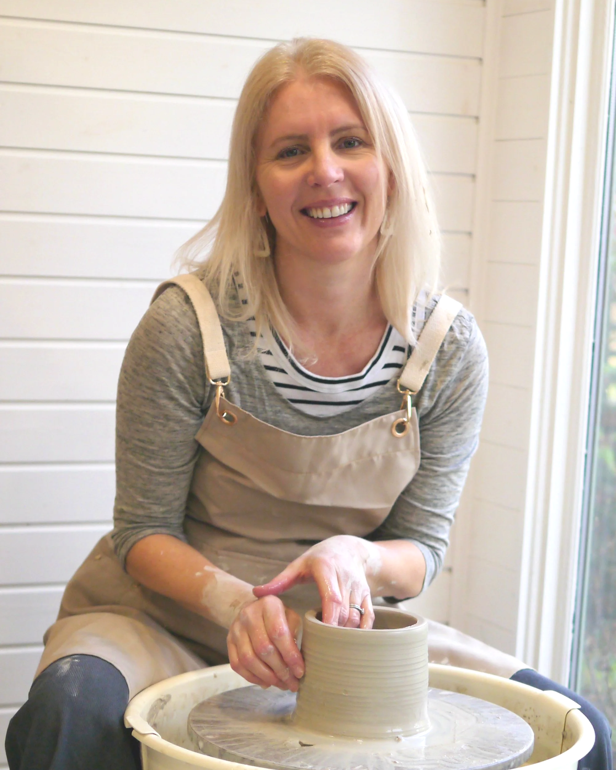 Anna Goss, potter at Anna Goss Ceramics, smiling while working on her pottery wheel, shaping clay into a pot with her hands, inside her pottery studio.