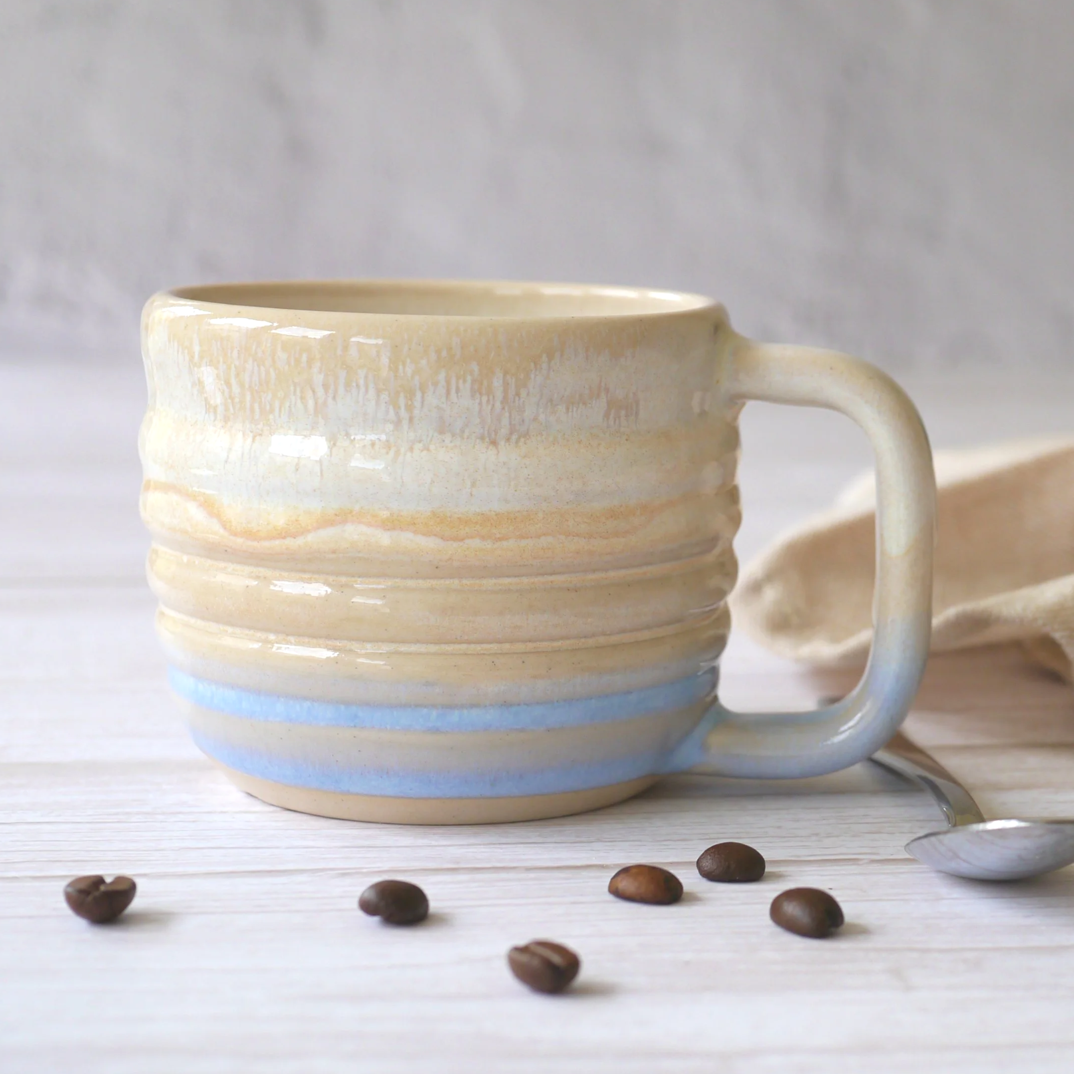 Hand-thrown stoneware mug. This mug features a beautiful blend of beige and light blue glazes over a rippled exterior, creating organic, flowing patterns that make every piece uniquely yours.