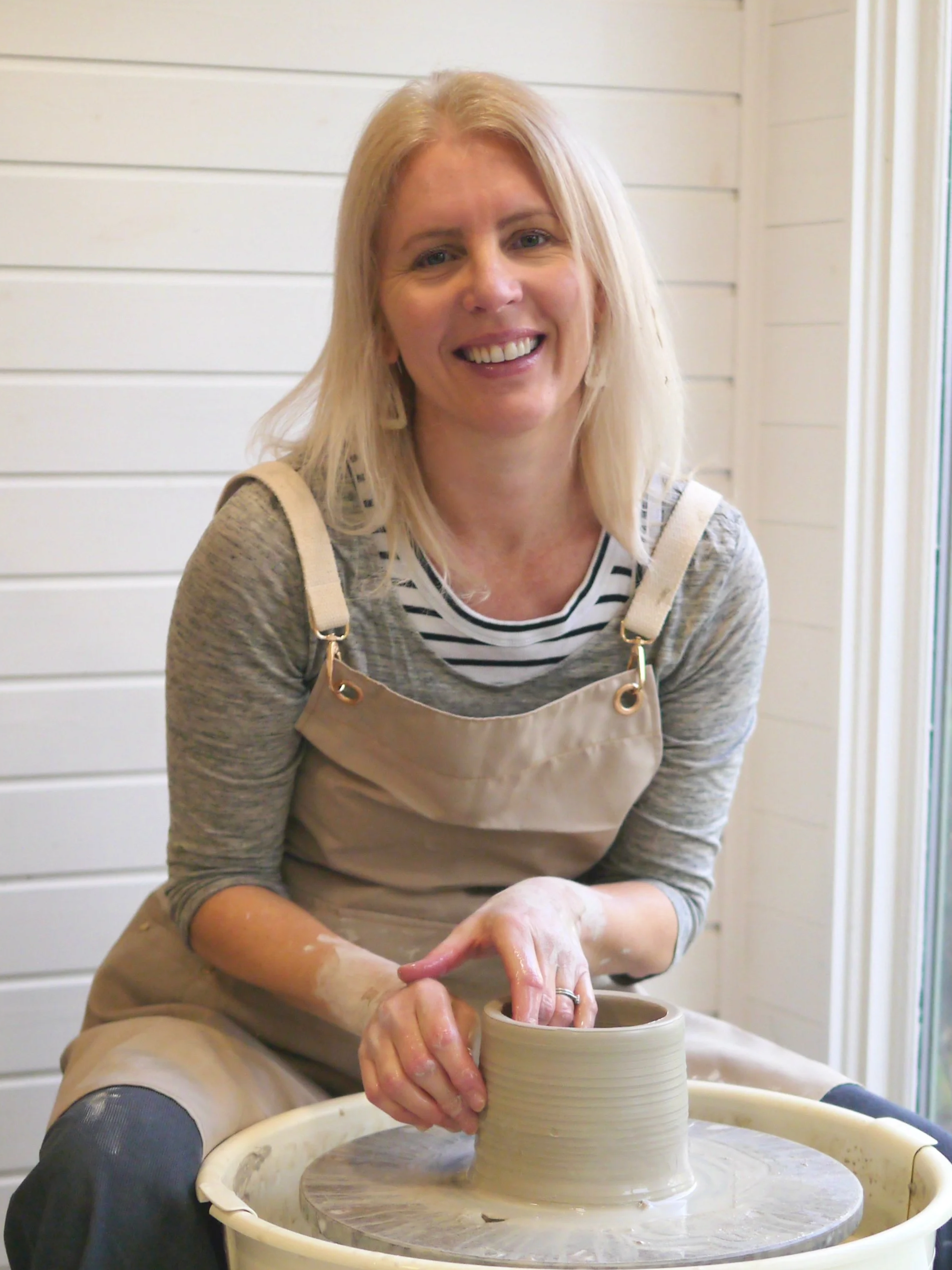 Anna Goss, potter at Anna Goss Ceramics, smiling while working on her pottery wheel, shaping clay into a pot with her hands, inside her pottery studio.