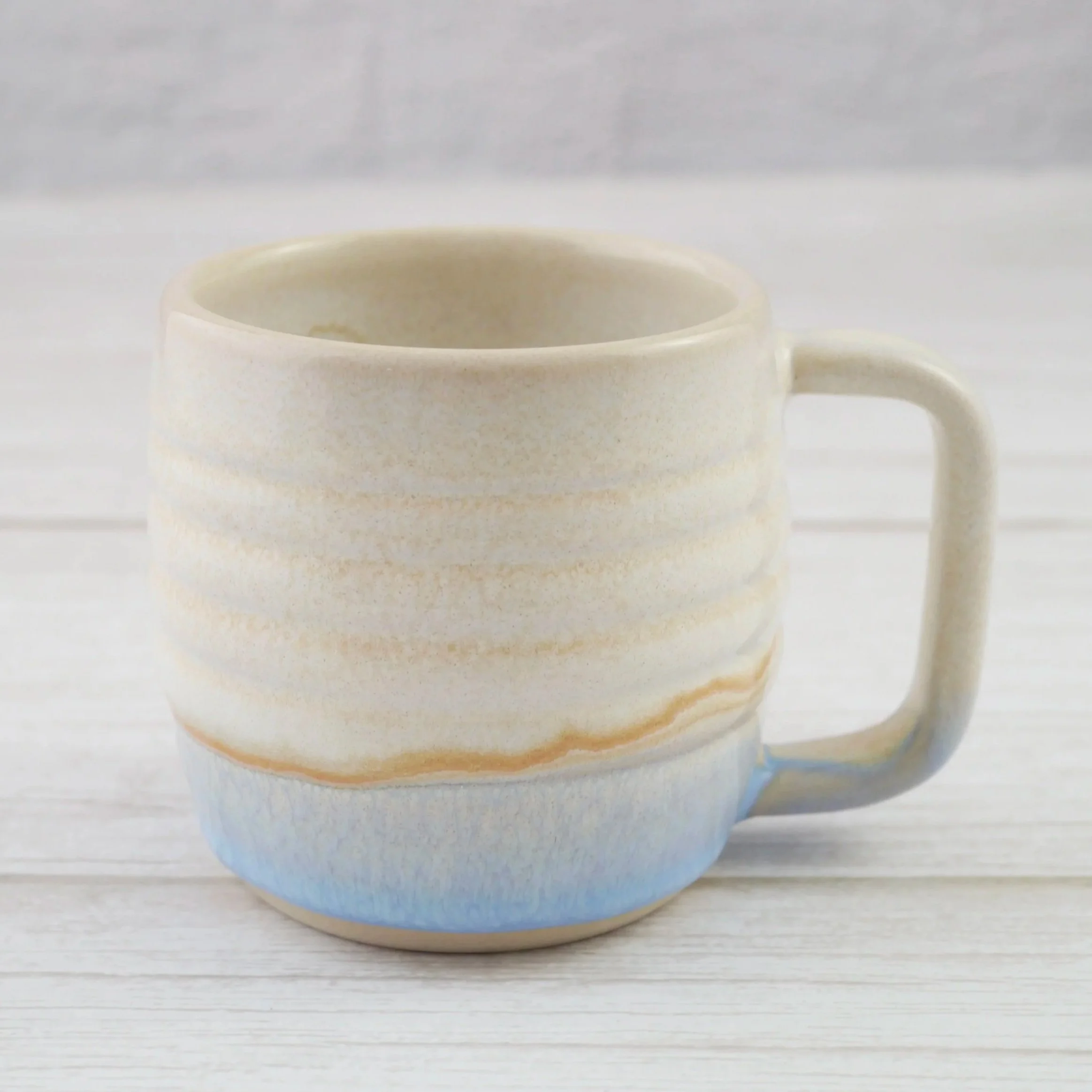 Hand-thrown stoneware mug. This mug features a beautiful blend of beige and light blue glazes over a rippled exterior, creating organic, flowing patterns that make every piece uniquely yours.