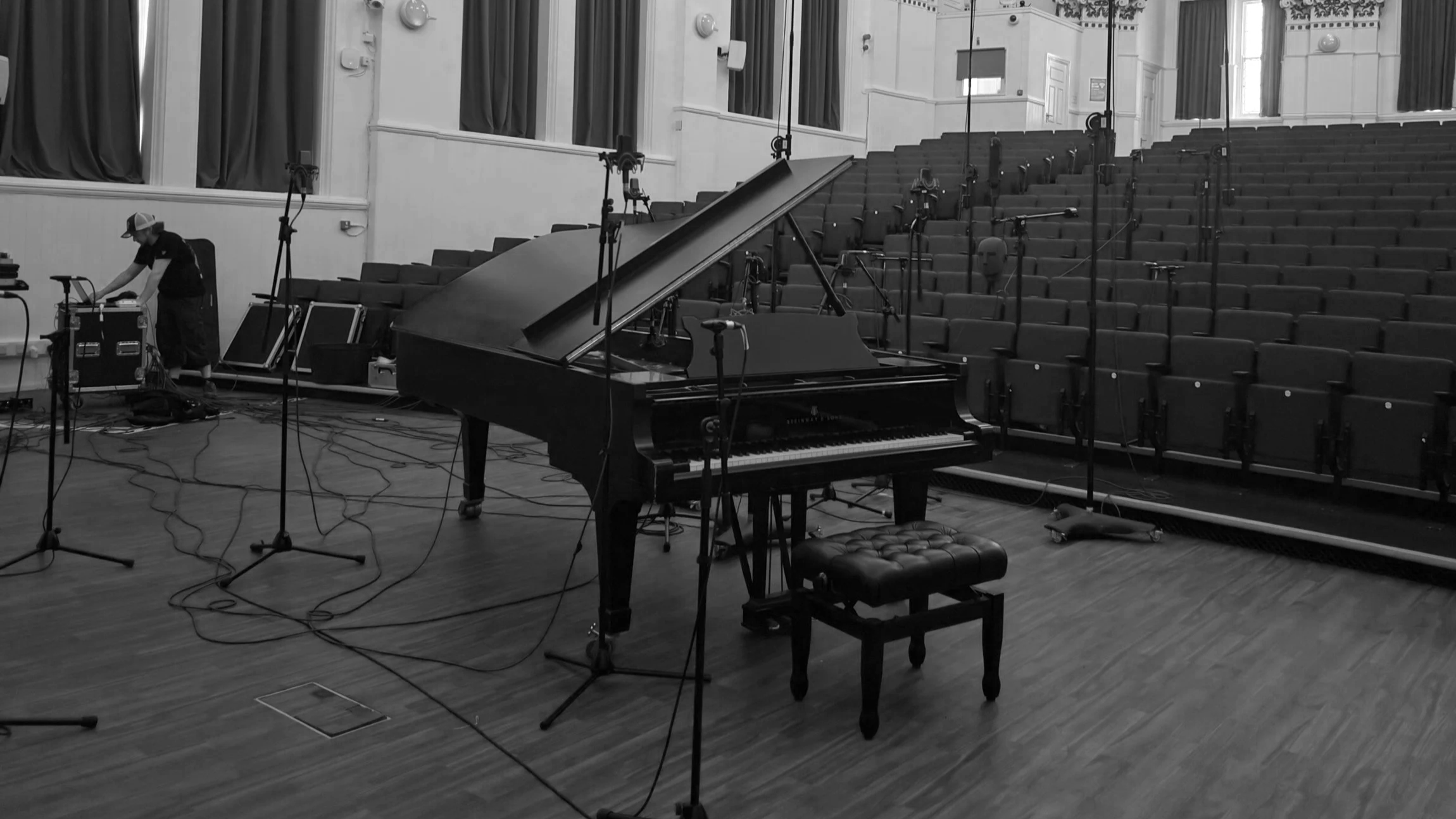 Empty concert hall with a grand piano in the center, surrounded by multiple microphones on stands, and an individual adjusting equipment on the left. The hall has tiered seating with dark curtains and large windows.