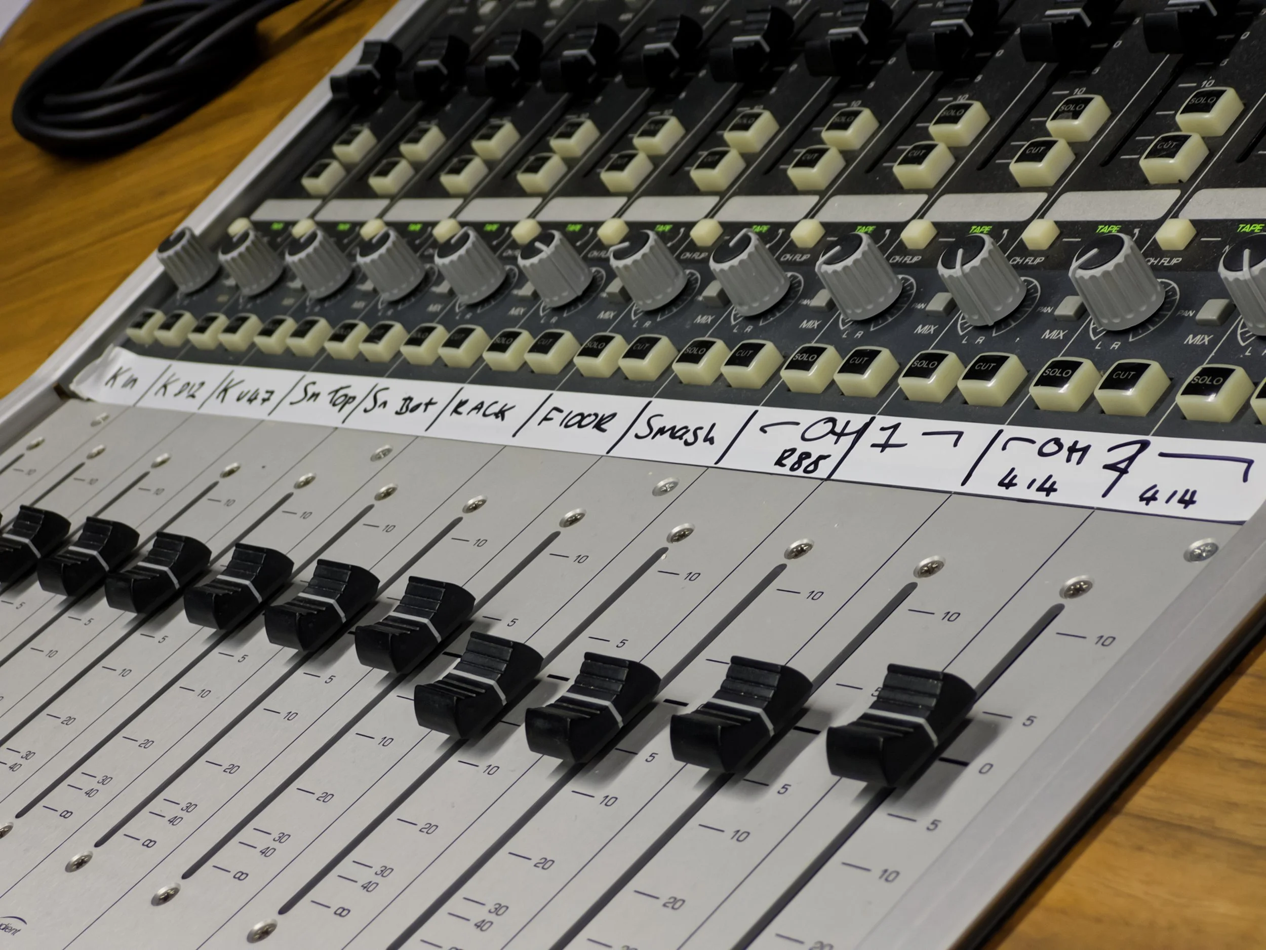 A sound mixing console with multiple sliders and knobs on a wooden surface, with handwritten labels on each channel.