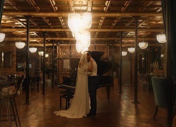A bride and groom sharing a kiss in a dimly lit, elegant venue with chandeliers and a grand piano.