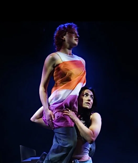 Performer, Alicia Meehan, holds Lucy Harries, who is sat on Alicia's shoulder. Lucy is wrapped in a lesbian flag.