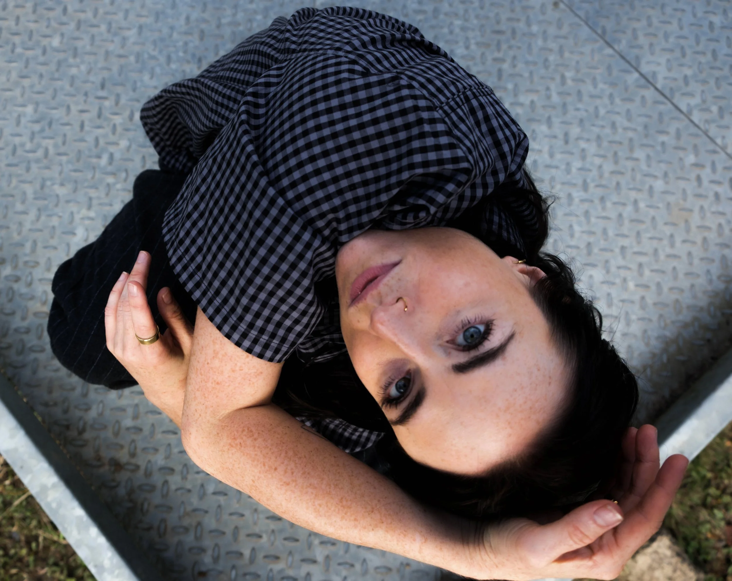 A woman lying on her back on a grey metal surface, with her head turned to the side and one eye open, looking at the camera. She has tattoos on her arm, freckles on her face, and is wearing a black and grey checkered shirt.