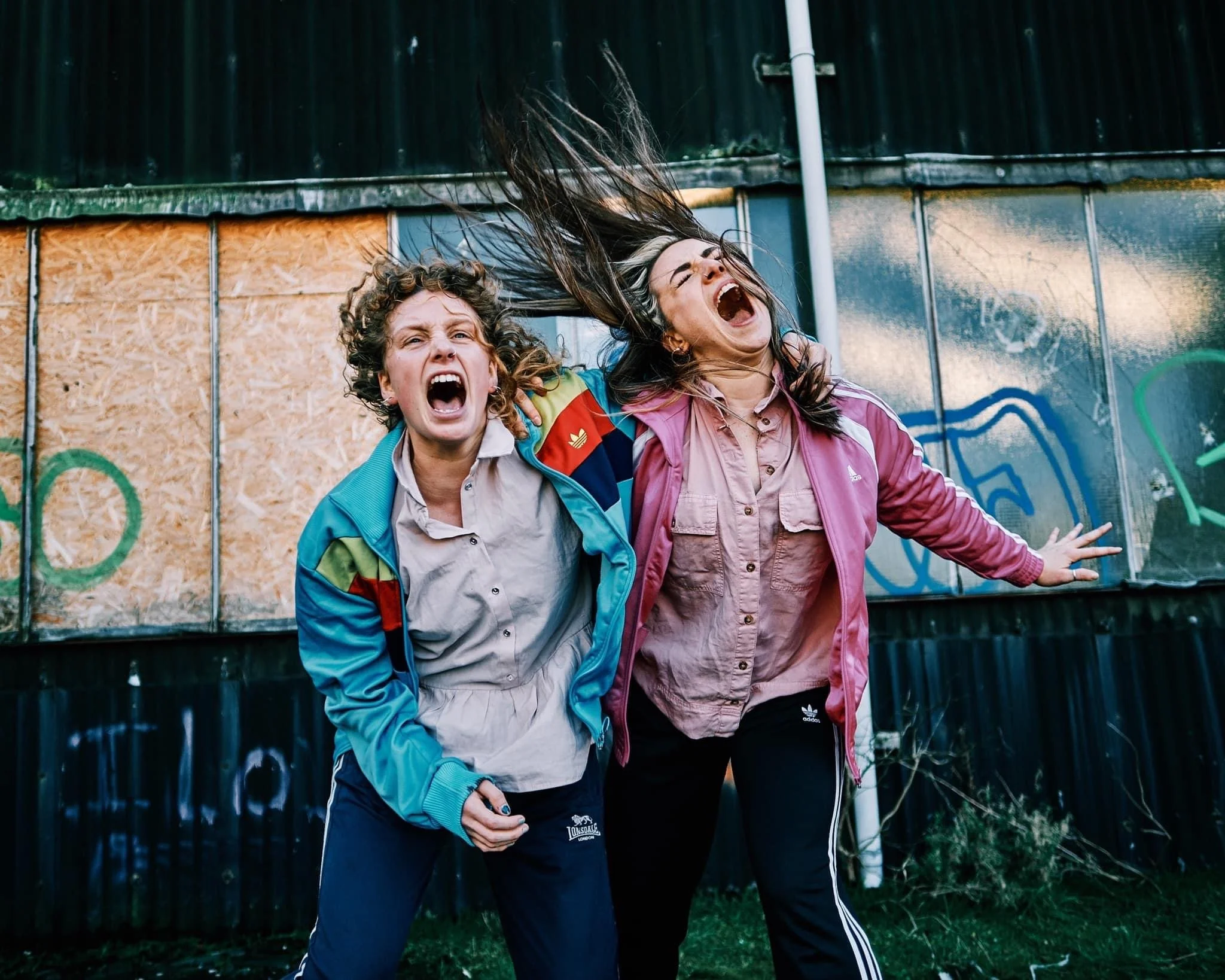 Image shows performers Alys North and Alicia Meehan dressed in pastel pinks and blues, screaming at the camera while standing in front of a graffitied building.