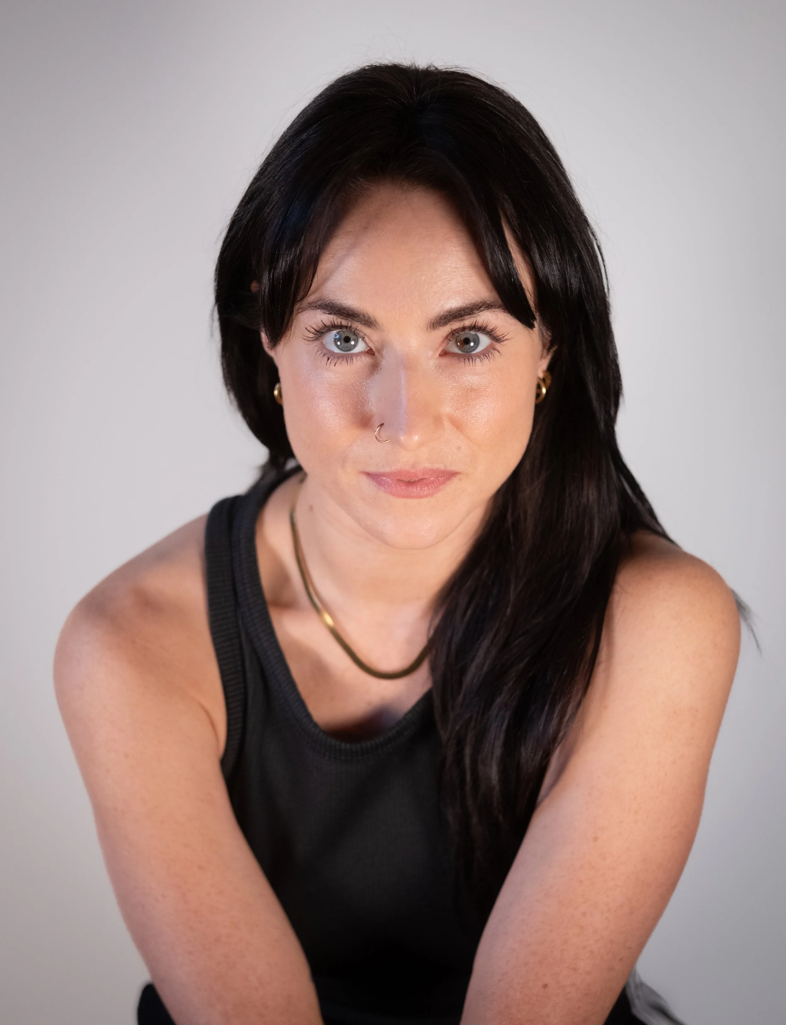 A headshot of Alicia. She/ they have long brown hair over one shoulder, looking piercingly at the camera. They are wearing a gold necklace, nose ring and earrings and are wearing a black vest.