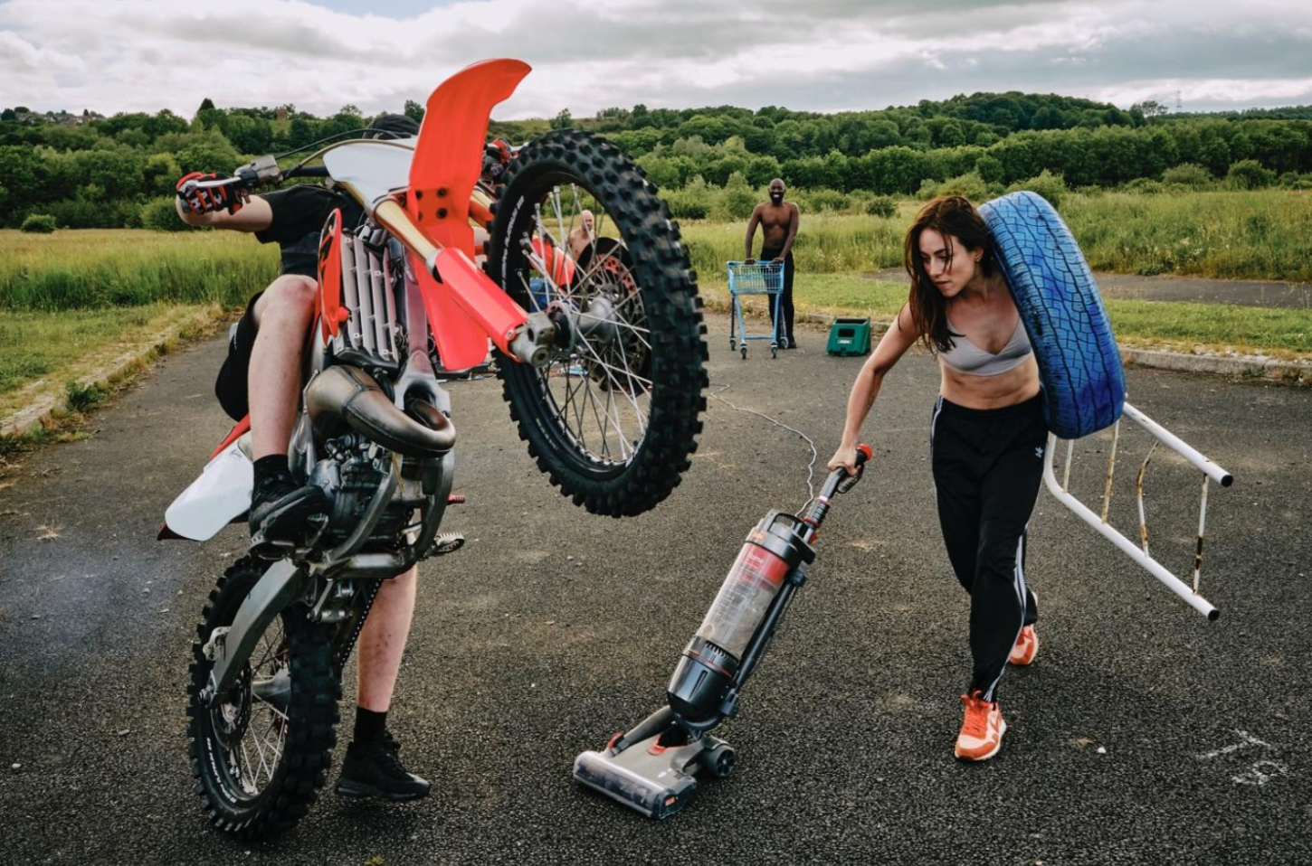 A woman in athletic clothes vacuuming the street while a motocross rider performs a wheelie on a dirt bike nearby. Two people are in the background, one with a shopping cart and another standing. Alicia Meehan - Movement Director and Co-Creator