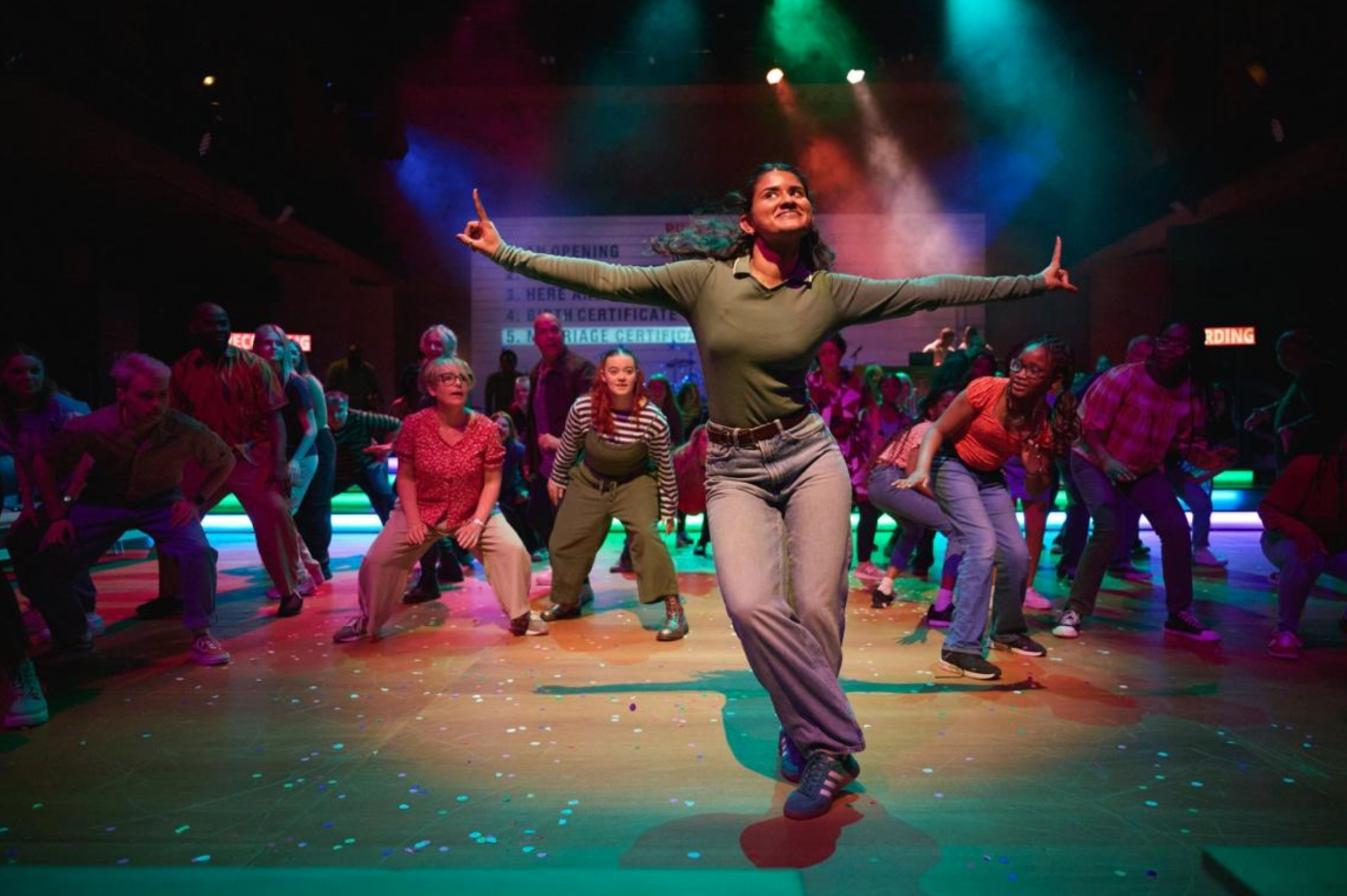 Image shows a performance of Public Record with a huge cast on stage, all paying attention to one performer telling a story. Alicia Meehan was Associate Choreographer.