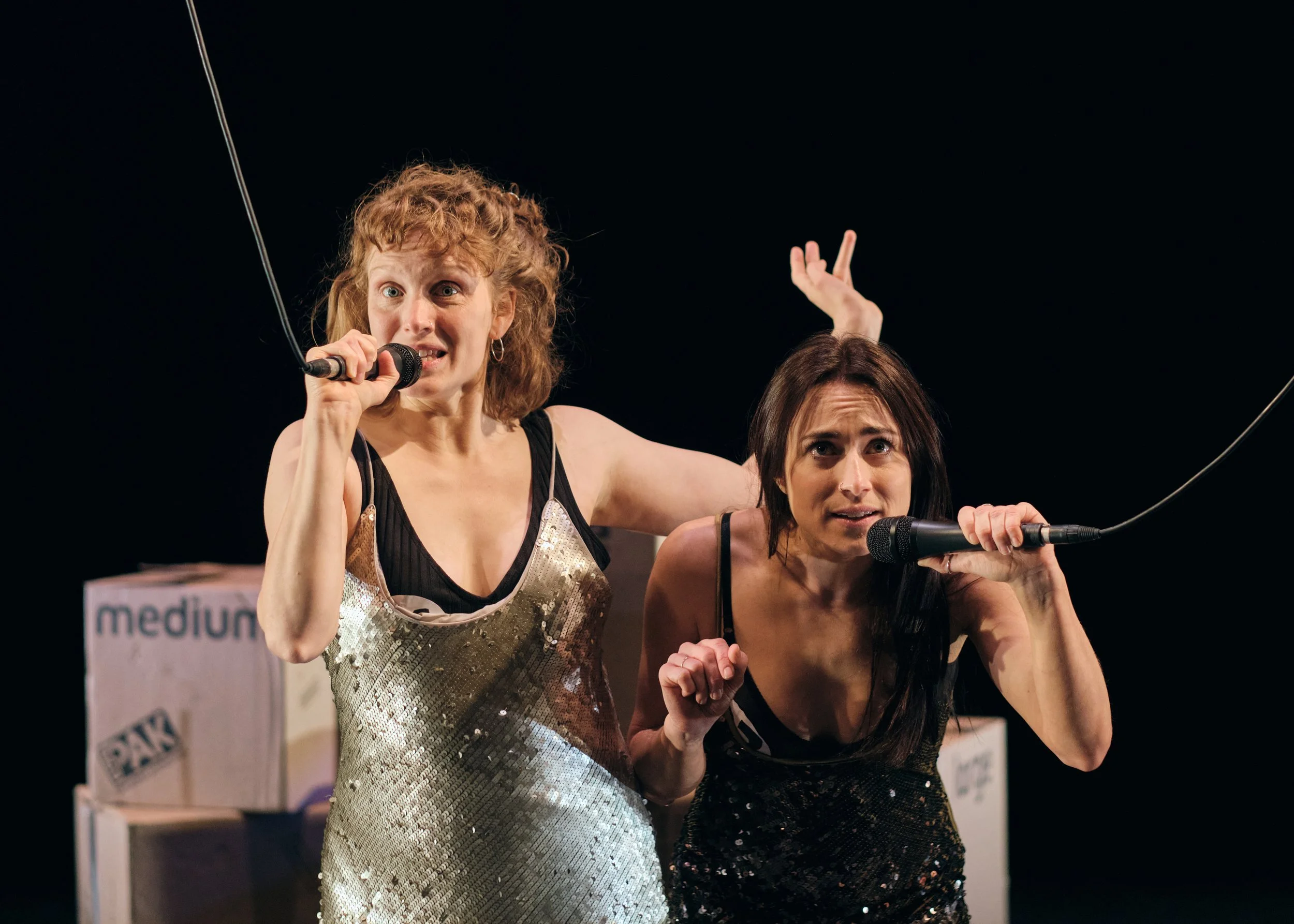 Two women singing or speaking into microphones on stage with a dark background and cardboard boxes behind them.