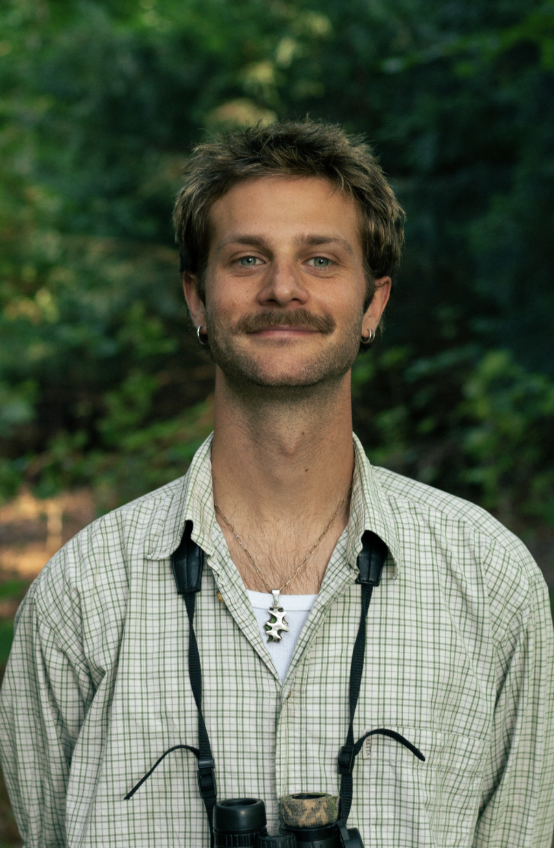A man with a mustache and earrings is standing outdoors in a wooded area, wearing a plaid shirt, with binoculars around his neck and a necklace, smiling at the camera.