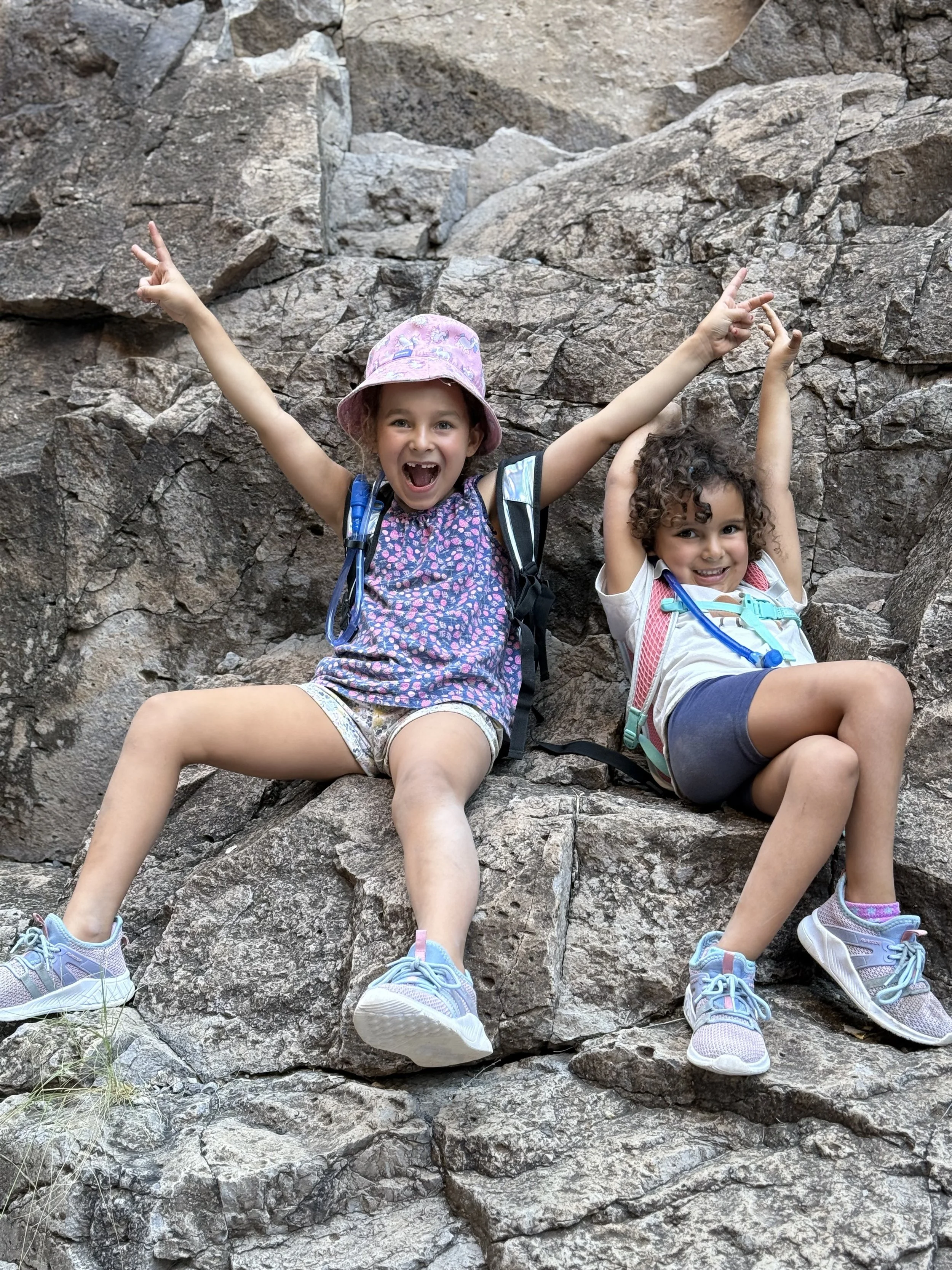 Two young girls sitting on a rocky mountain ledge with arms raised and making peace signs. They are wearing casual summer clothes, backpacks, and sneakers, and smiling happily.