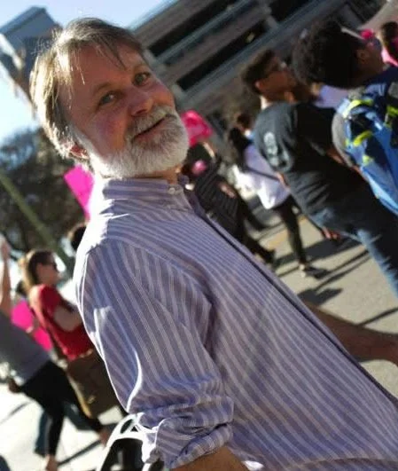 A man with white hair and beard smiling outdoors in a crowd.