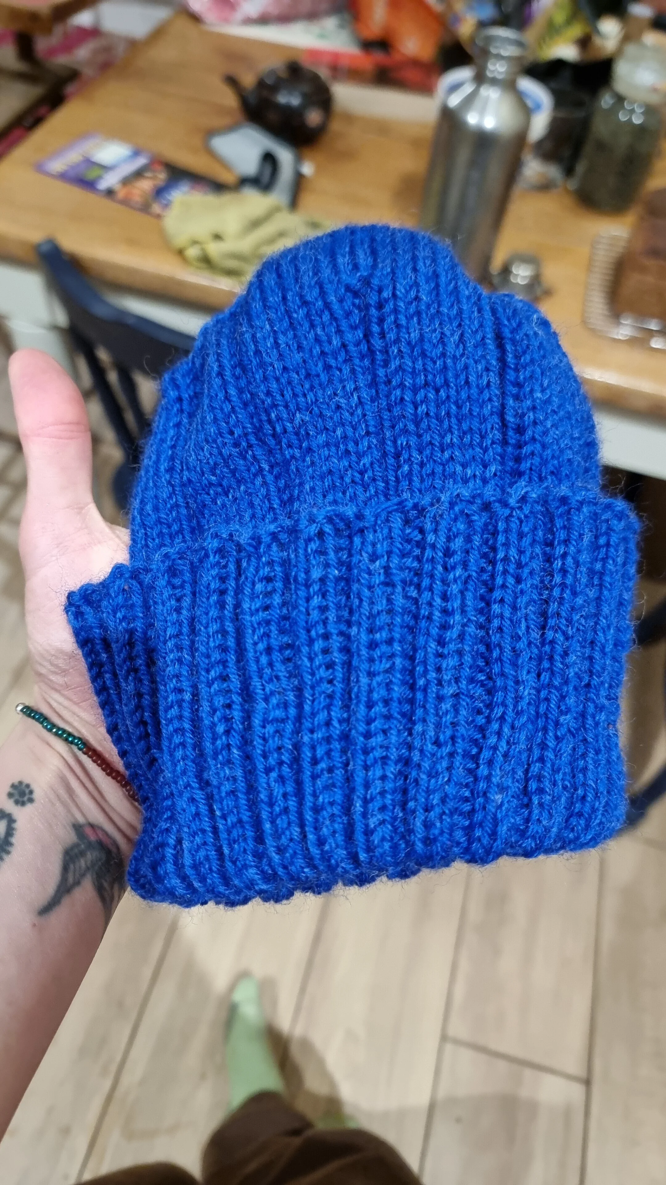 A hand holding a blue knit beanie in a kitchen with a wooden table and various bottles and objects on it in the background.