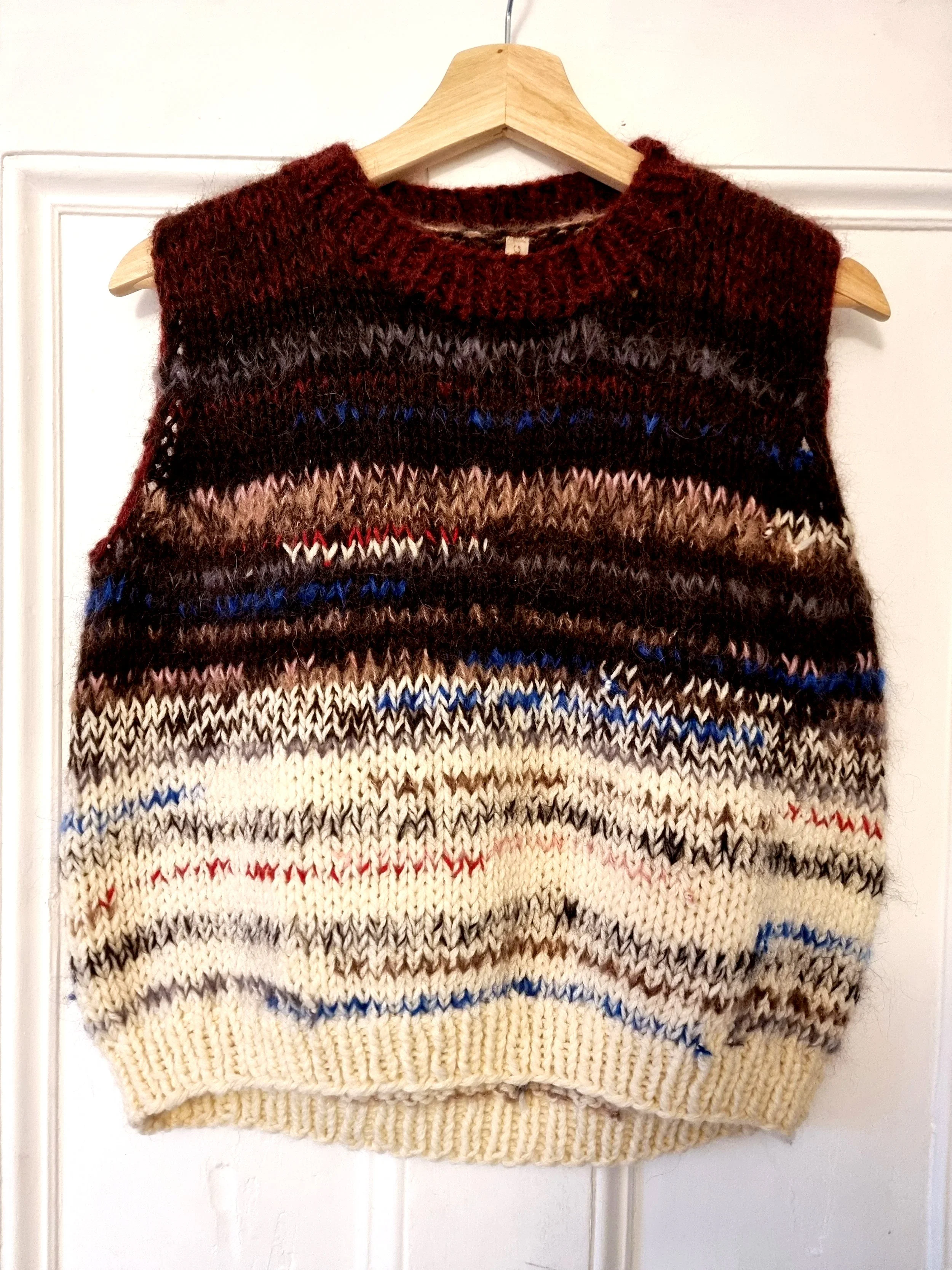 Colorful knitted sleeveless sweater with horizontal stripes in shades of brown, black, cream, red, and blue hanging on a wooden hanger.