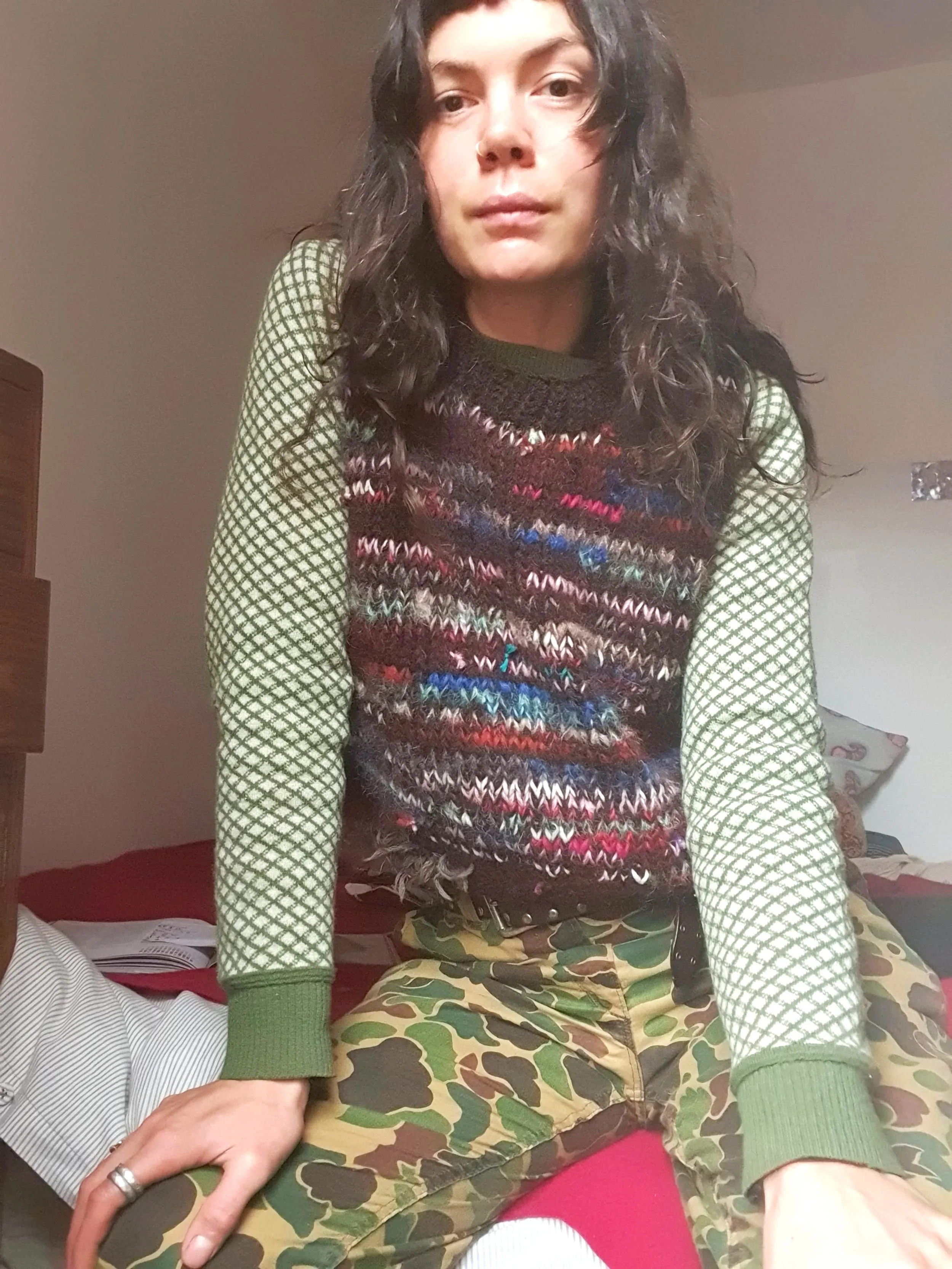 A person with long curly hair, wearing a green and black patterned sweater and camouflage pants, sitting on a bed in a room with a beige wall.