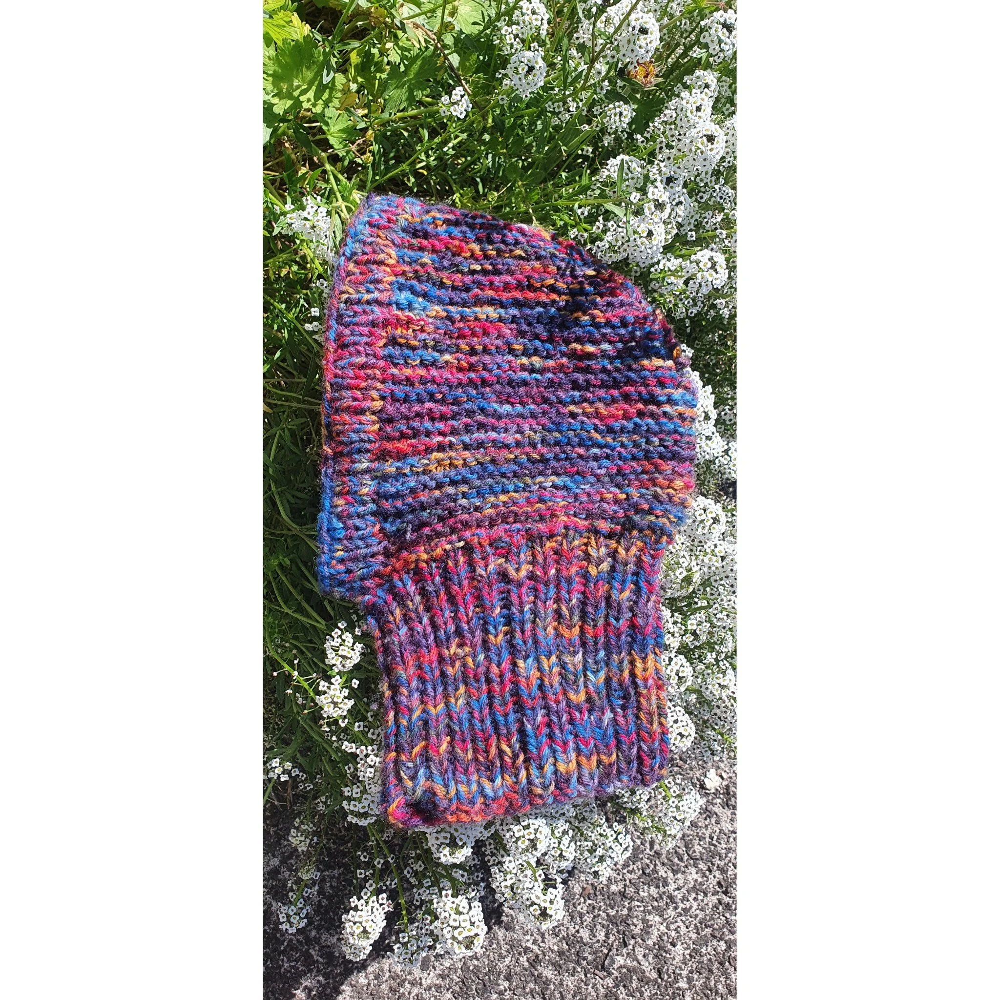 A colorful knitted hat resting on white flowers and green foliage.