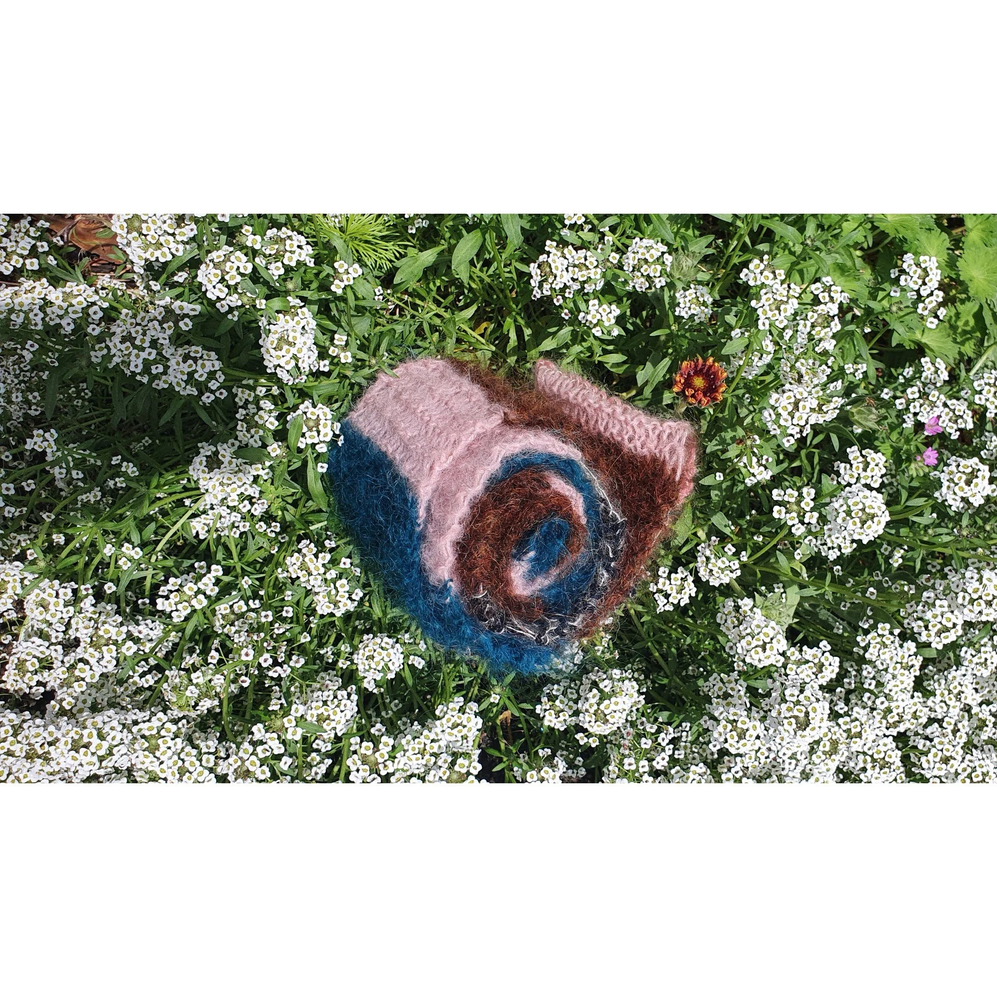 Multicoloured scarf rolled into a spiral resting on a bed of white flowers with some greenery and a single dark orange flower.