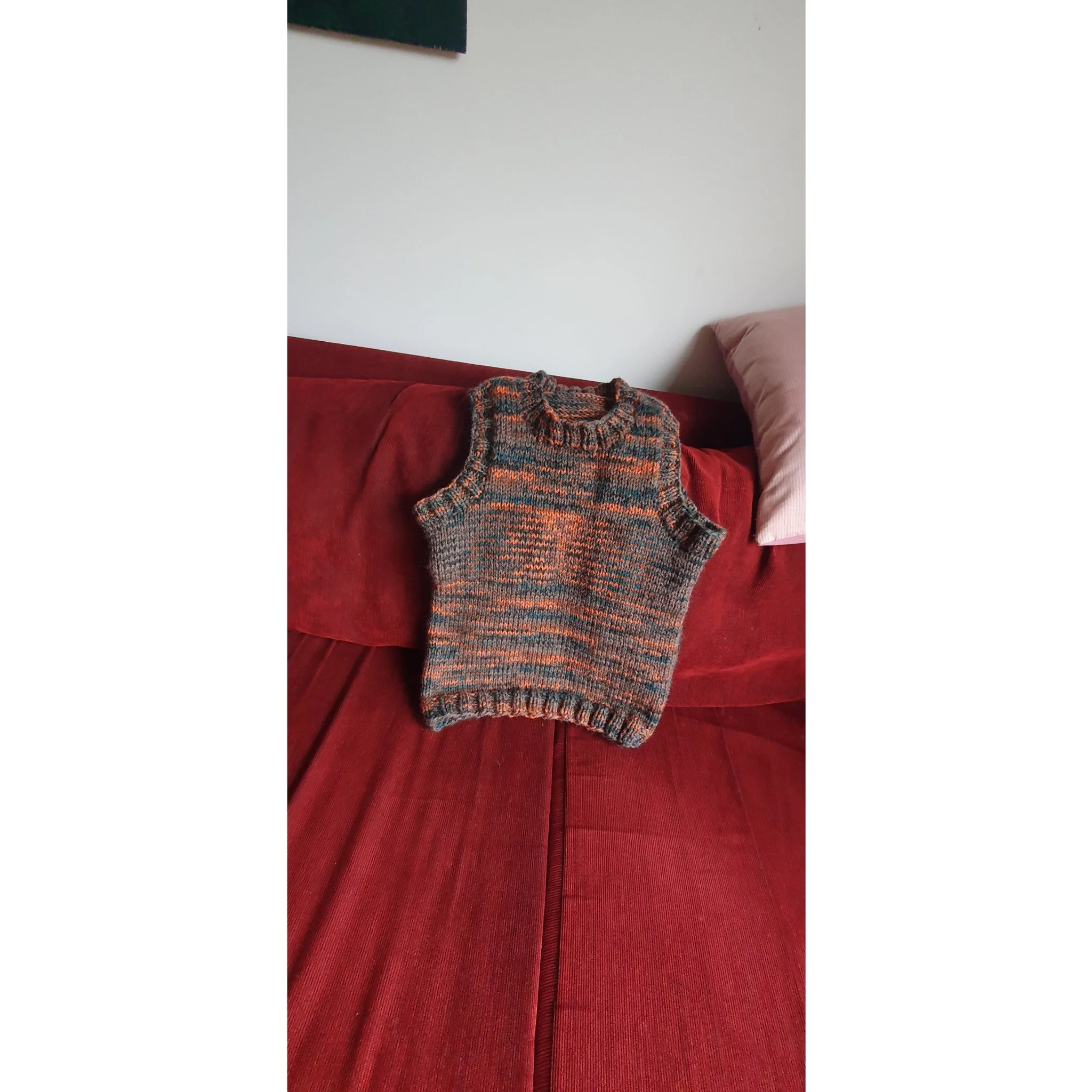 A multicolored knitted vest lying on a red sofa with a pink pillow nearby.