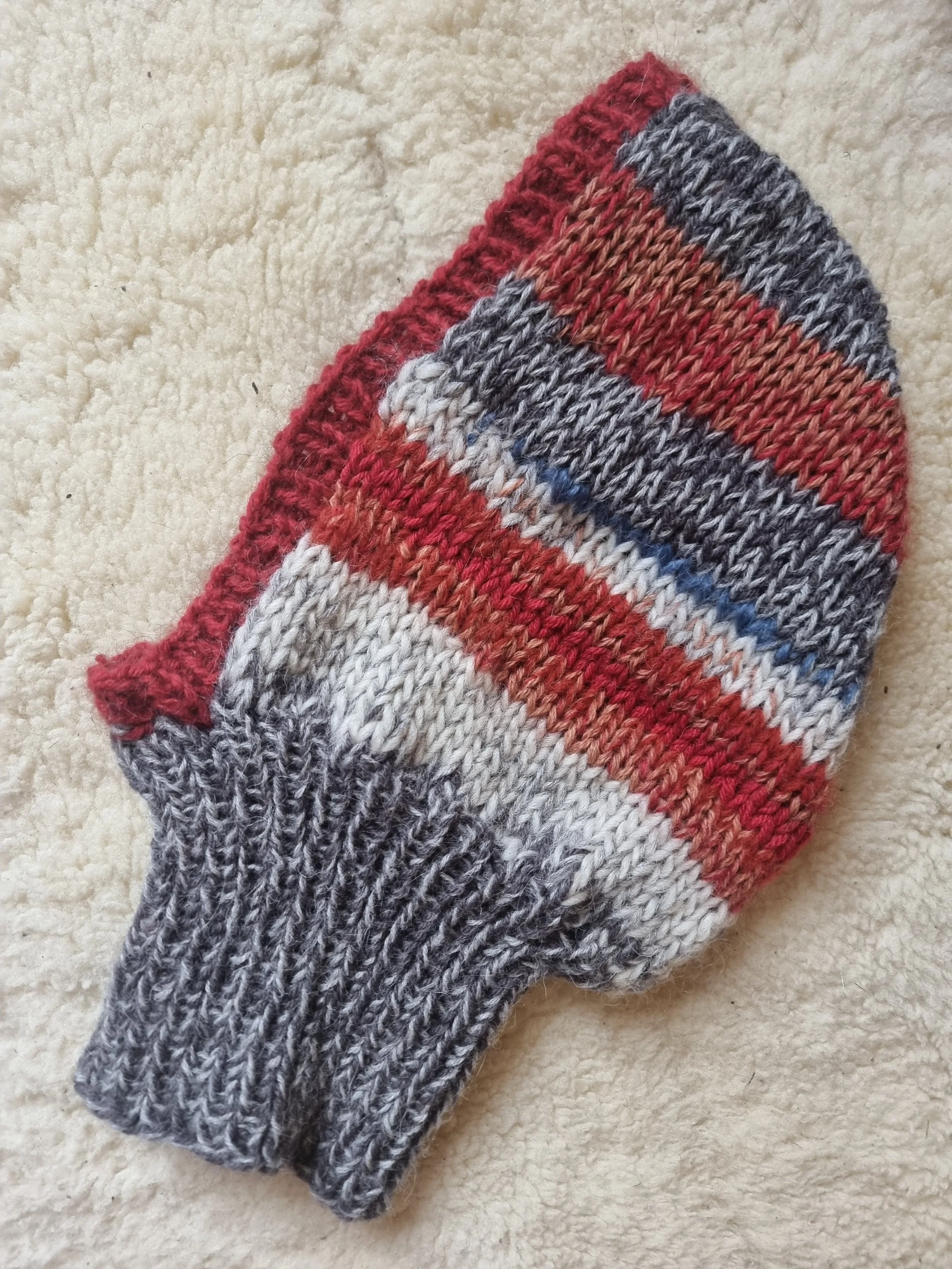 A knitted winter balaclava with red, gray, white, and black stripes placed on a beige, fuzzy surface.