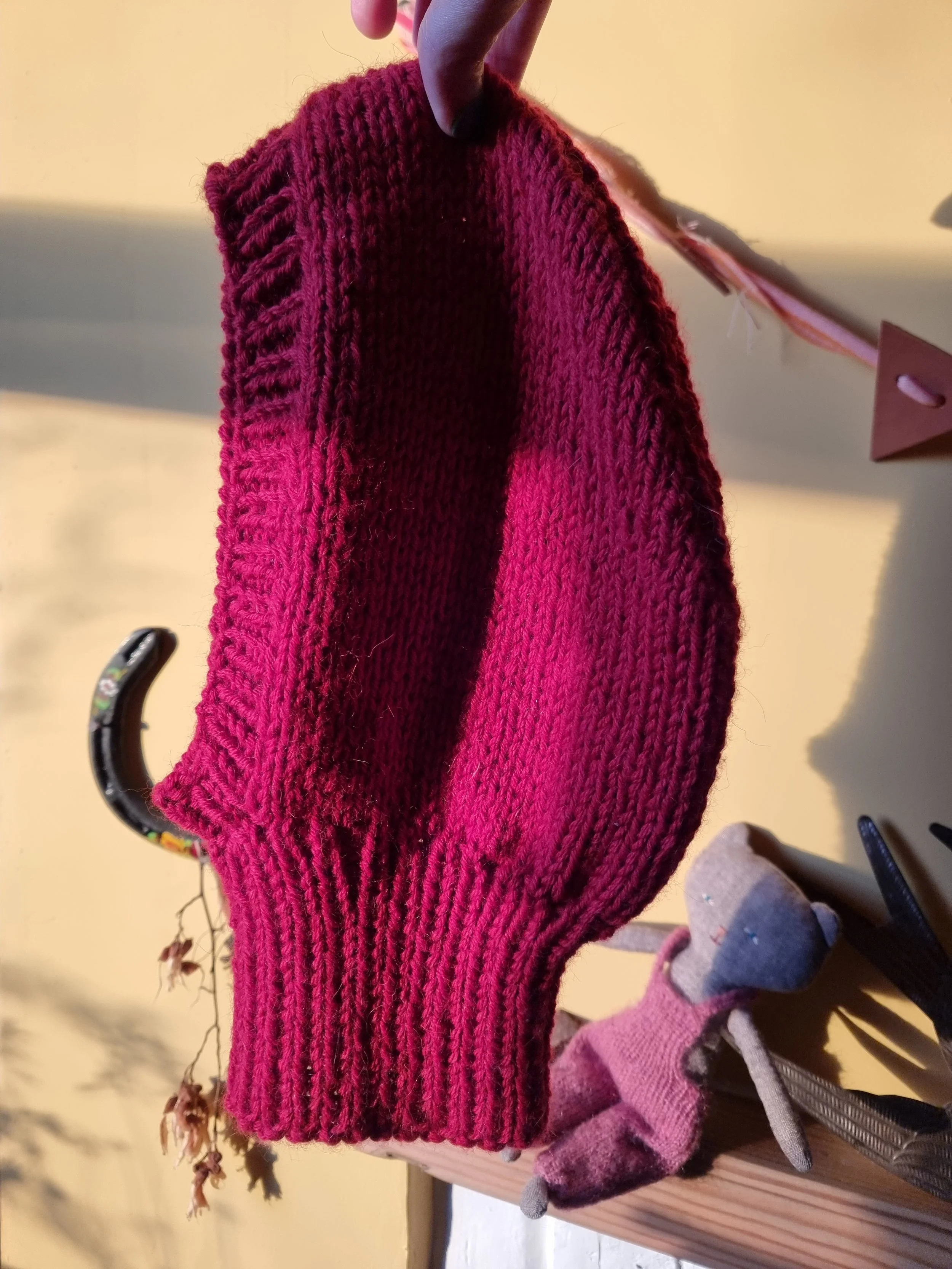 Close-up of a knitted red balaclava being held up by a hand against a yellow background near a clothes hook.