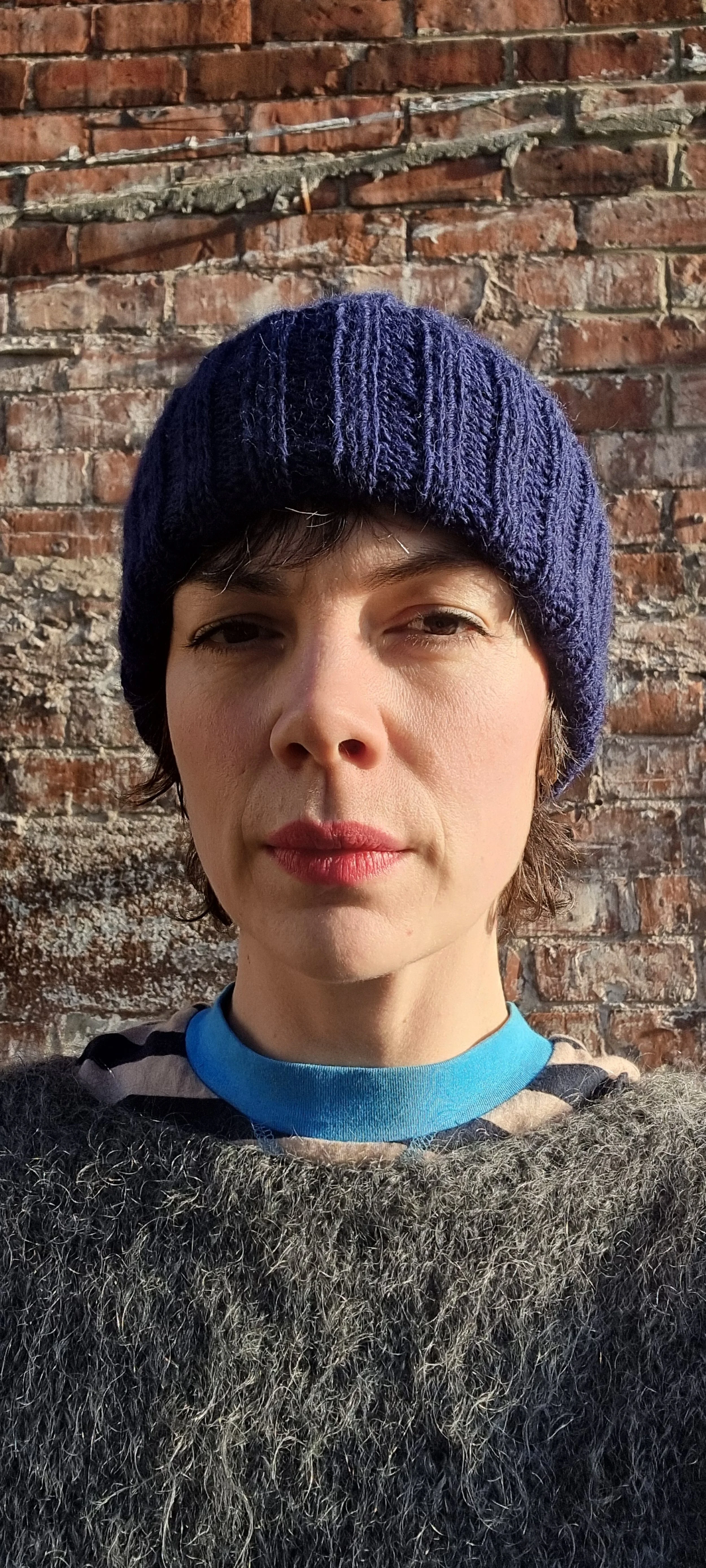 Close-up of a woman wearing a navy knit beanie, multi-colored striped sweater, and a cooler blue shirt underneath, standing in front of a weathered brick wall.