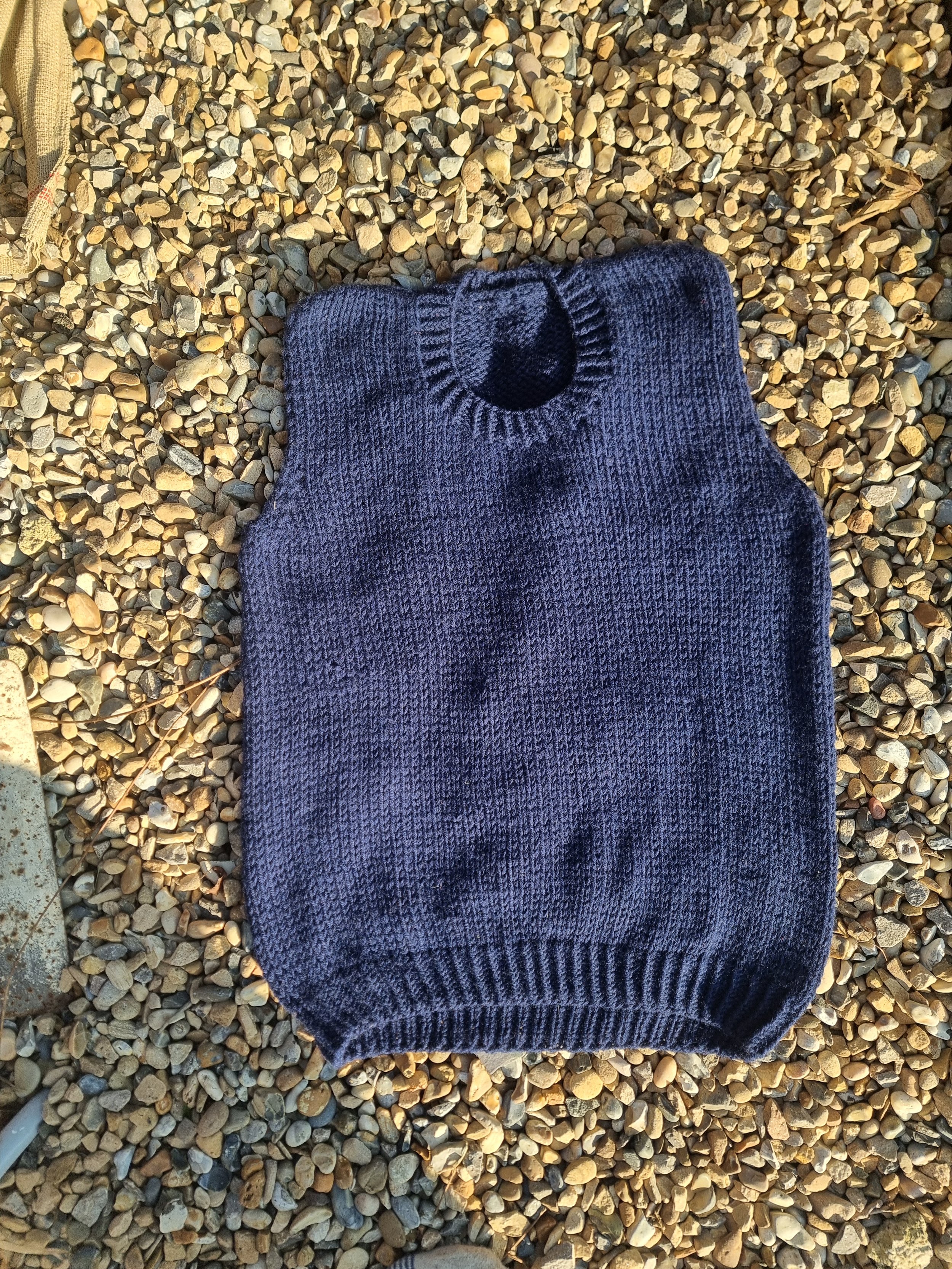 A navy blue knitted sweater with a blue collar, laid flat on a bed of small, tan and gray pebbles outdoors.