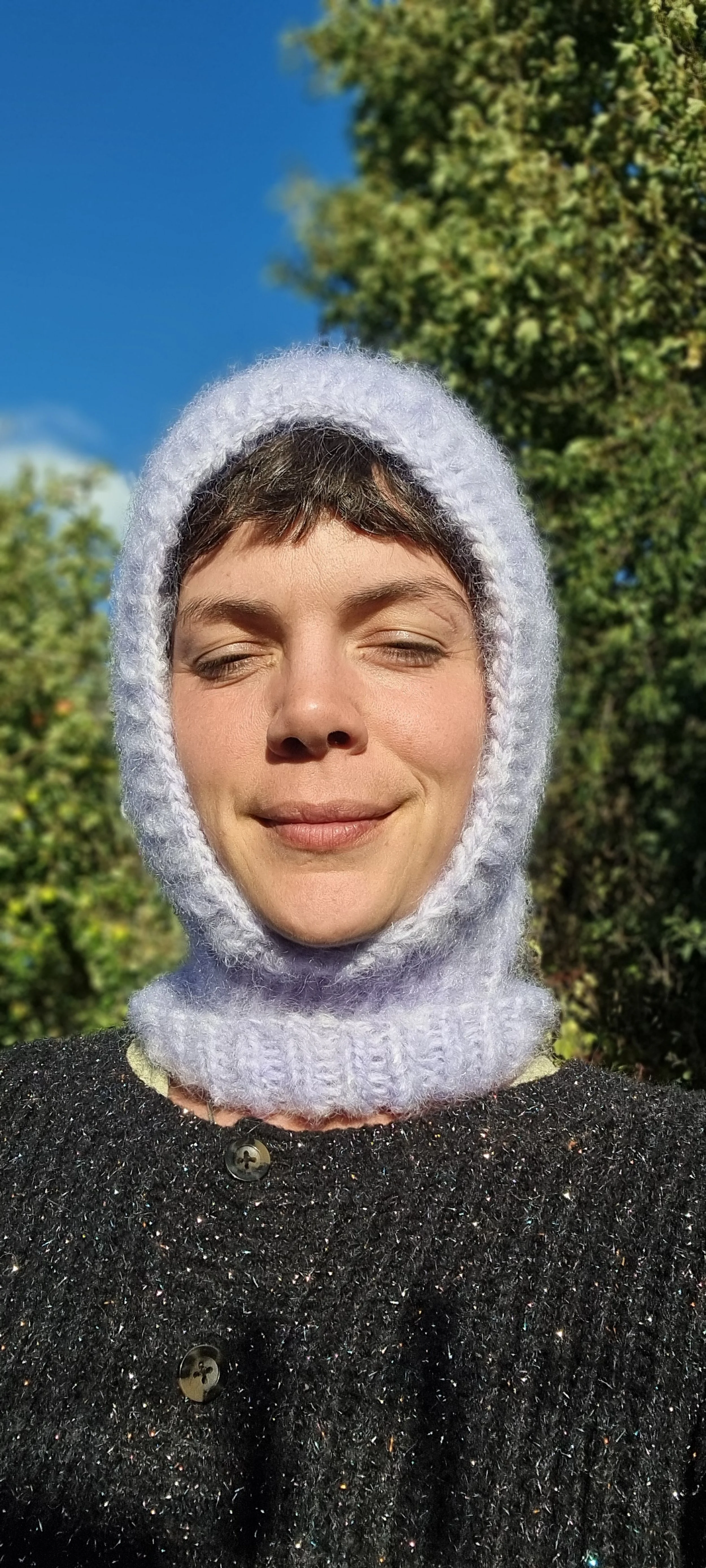 A woman is outdoors with her eyes closed, wearing a lilac knitted balaclava and a black, sparkly textured coat, with trees and a blue sky in the background.