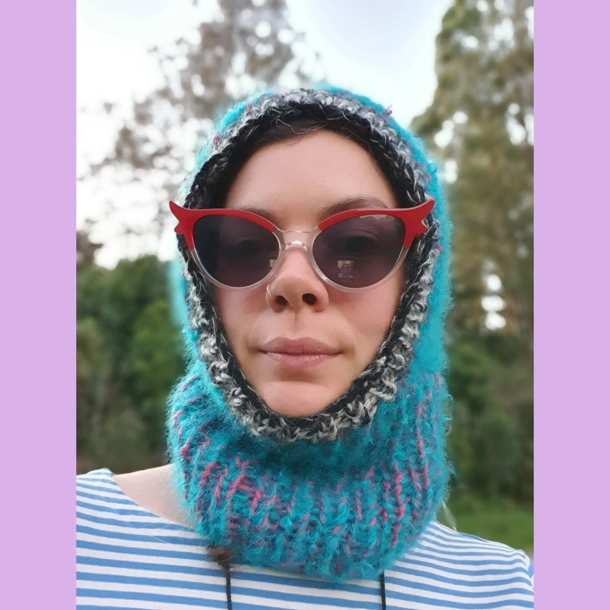 Person outdoors wearing round sunglasses, a colorful knitted balaclava and a striped shirt.
