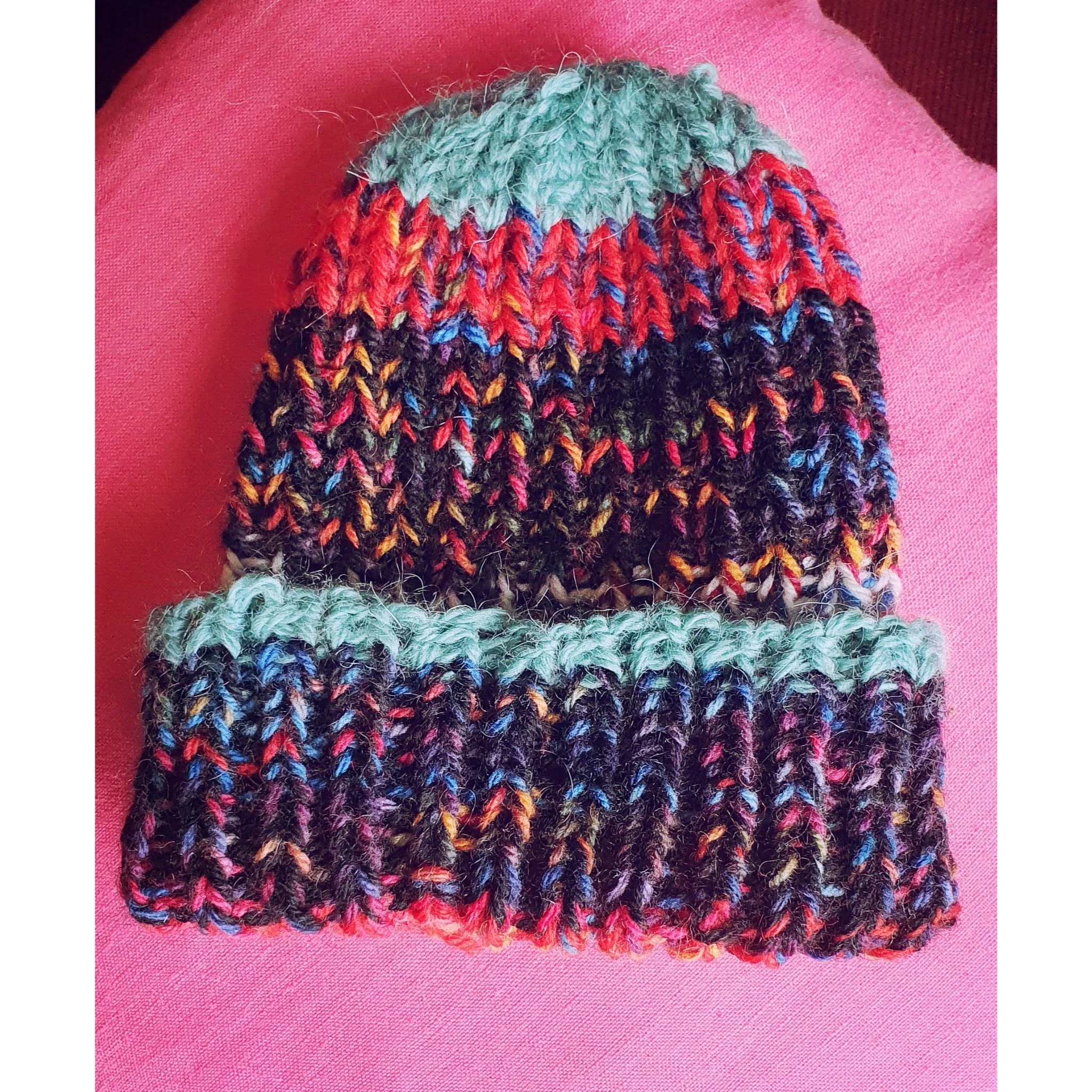 A colorful knit beanie hat with a folded brim, placed on a pink fabric background.