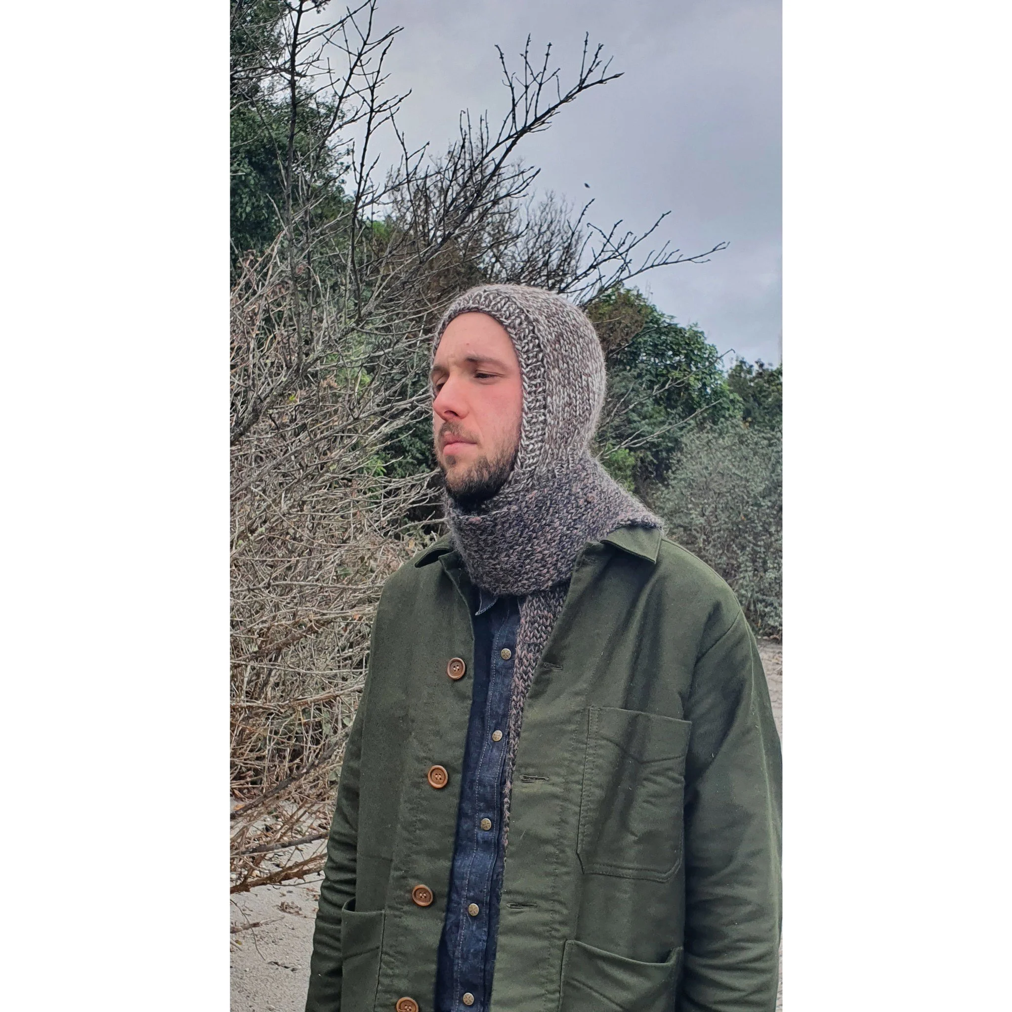 A man wearing a knit balaclava and a green jacket standing outdoors with bare trees and cloudy sky in the background.