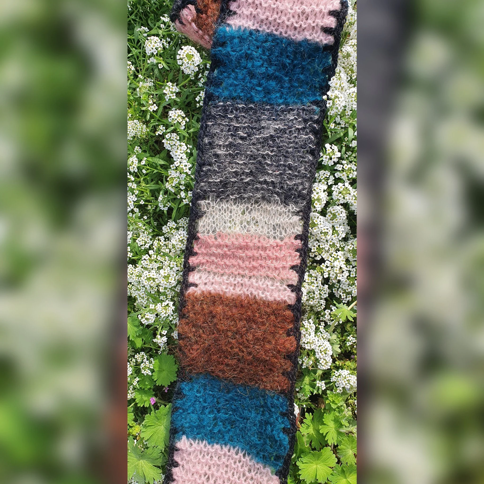 Colorful knitted scarf with horizontal stripes laid on green grass with small white flowers.