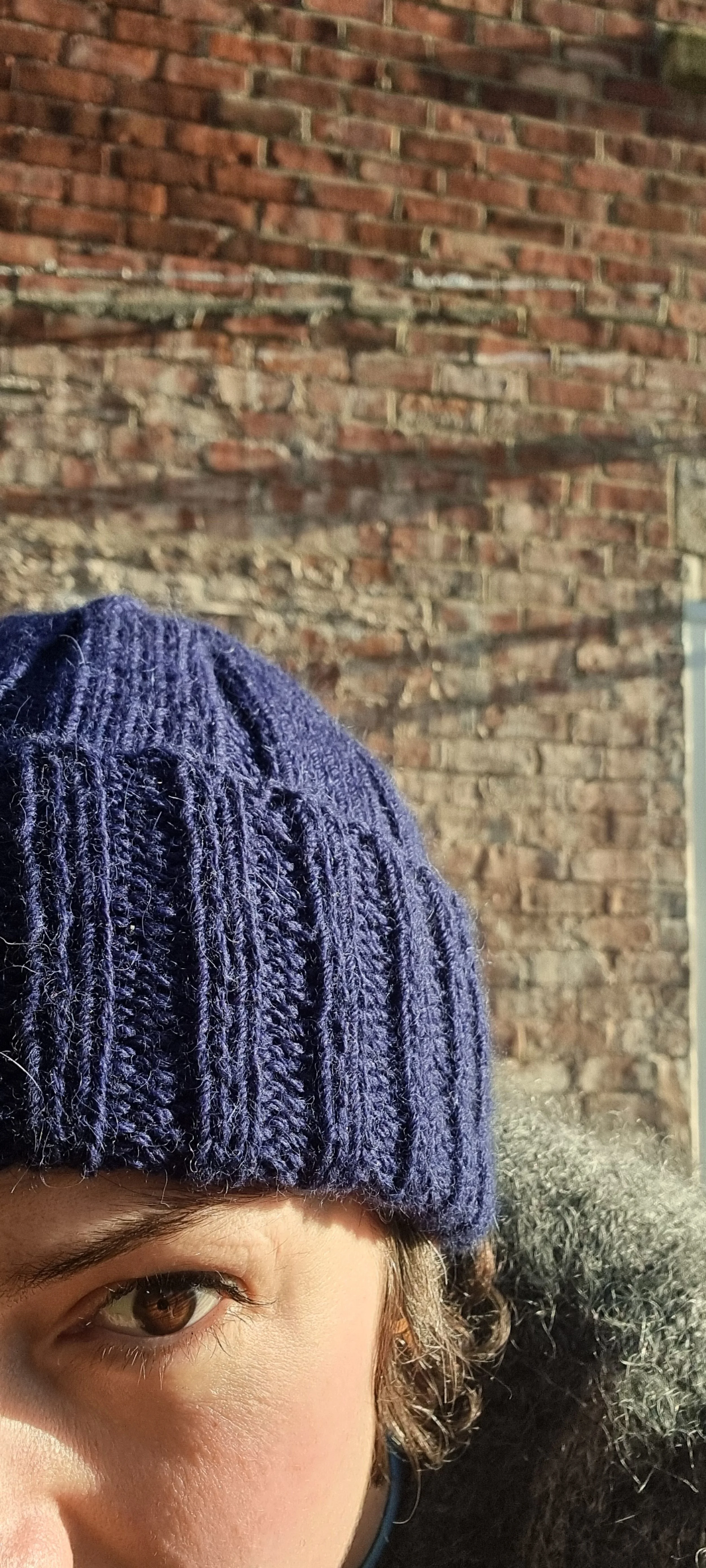 Close-up of a person wearing a navy knit beanie, showing part of their face including an eye, with a brick wall in the background.