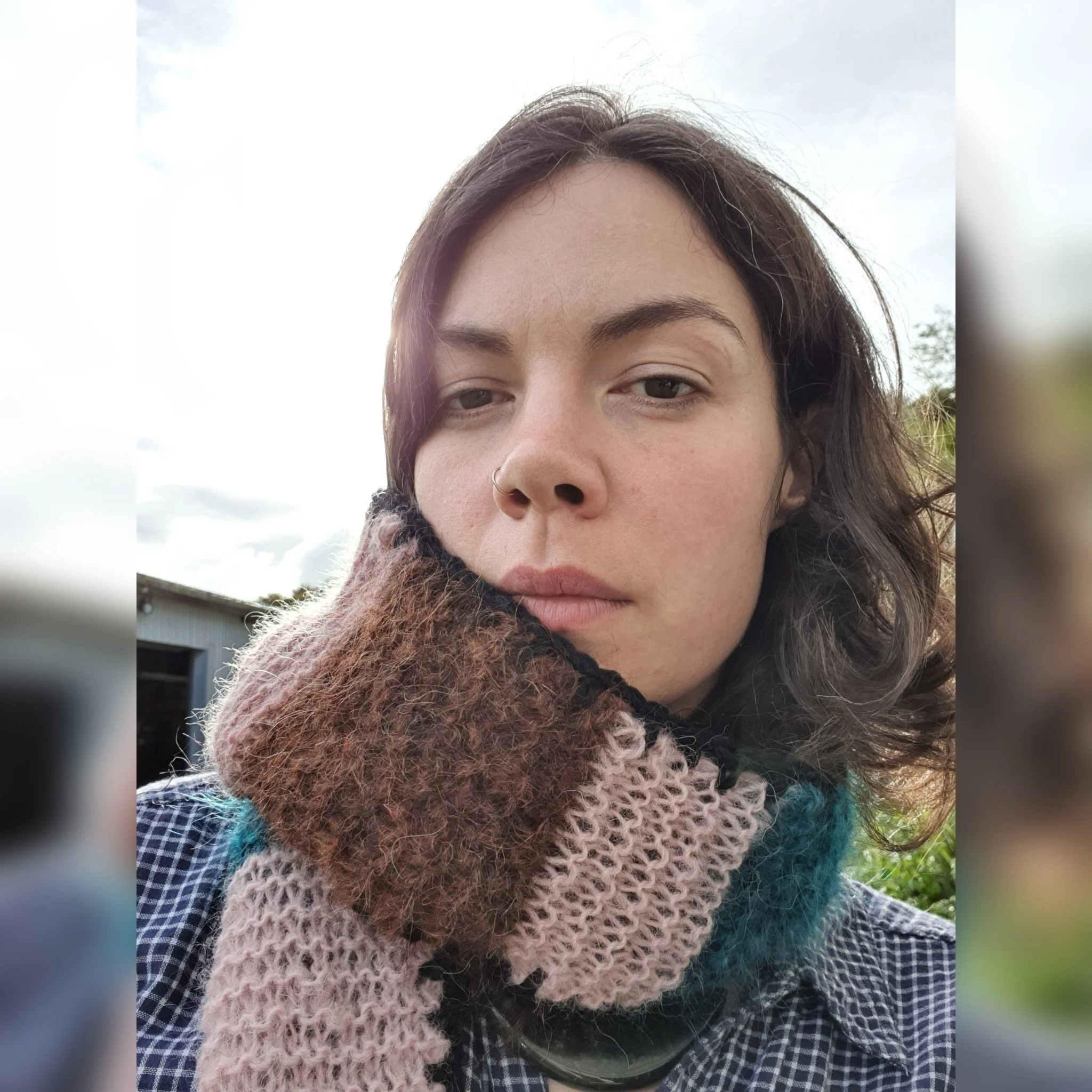 Close-up of a woman with dark wavy hair, wearing a checkered shirt and a multi-colored knitted scarf, standing outdoors with cloudy sky in the background.