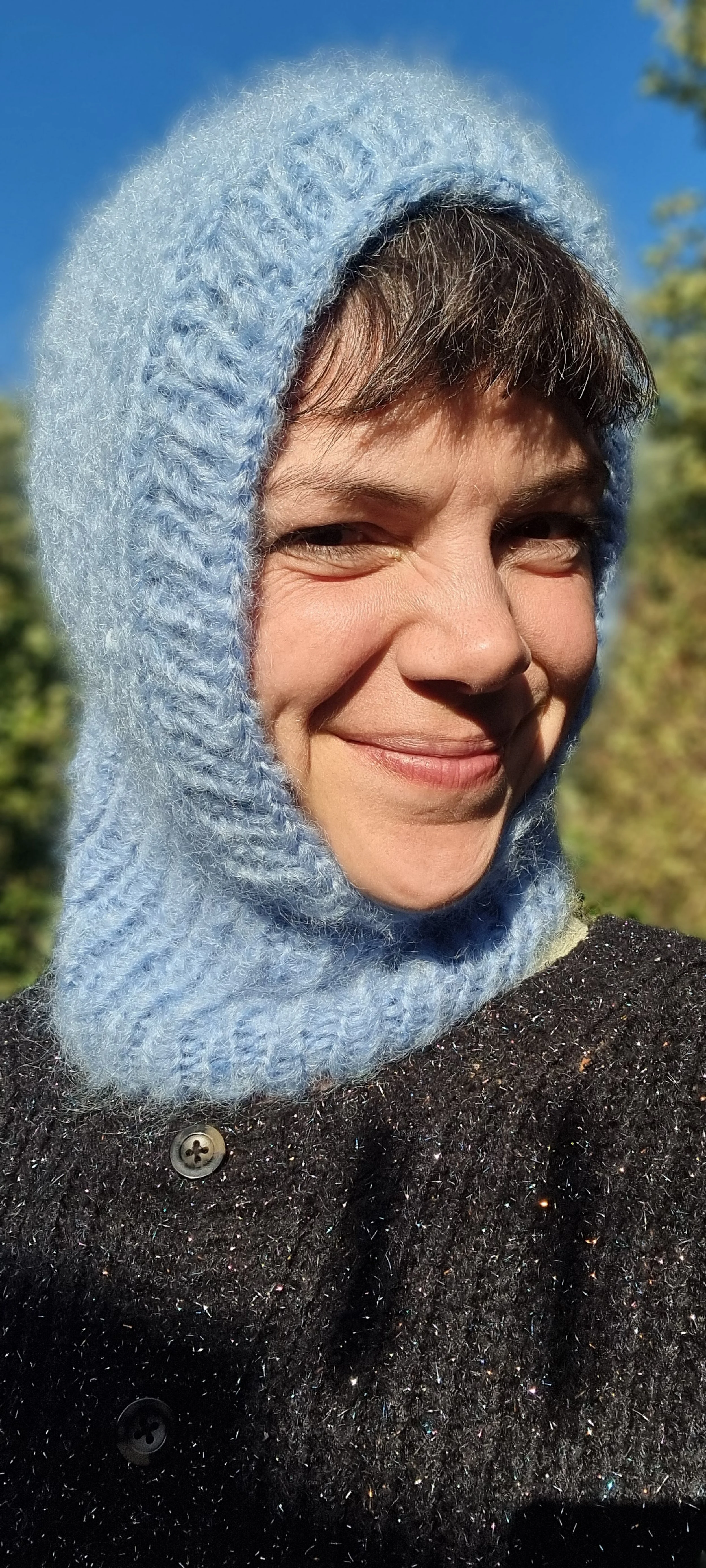 A woman smiling outdoors in sunlight, wearing a light blue knit balaclava and a dark, sparkly coat.