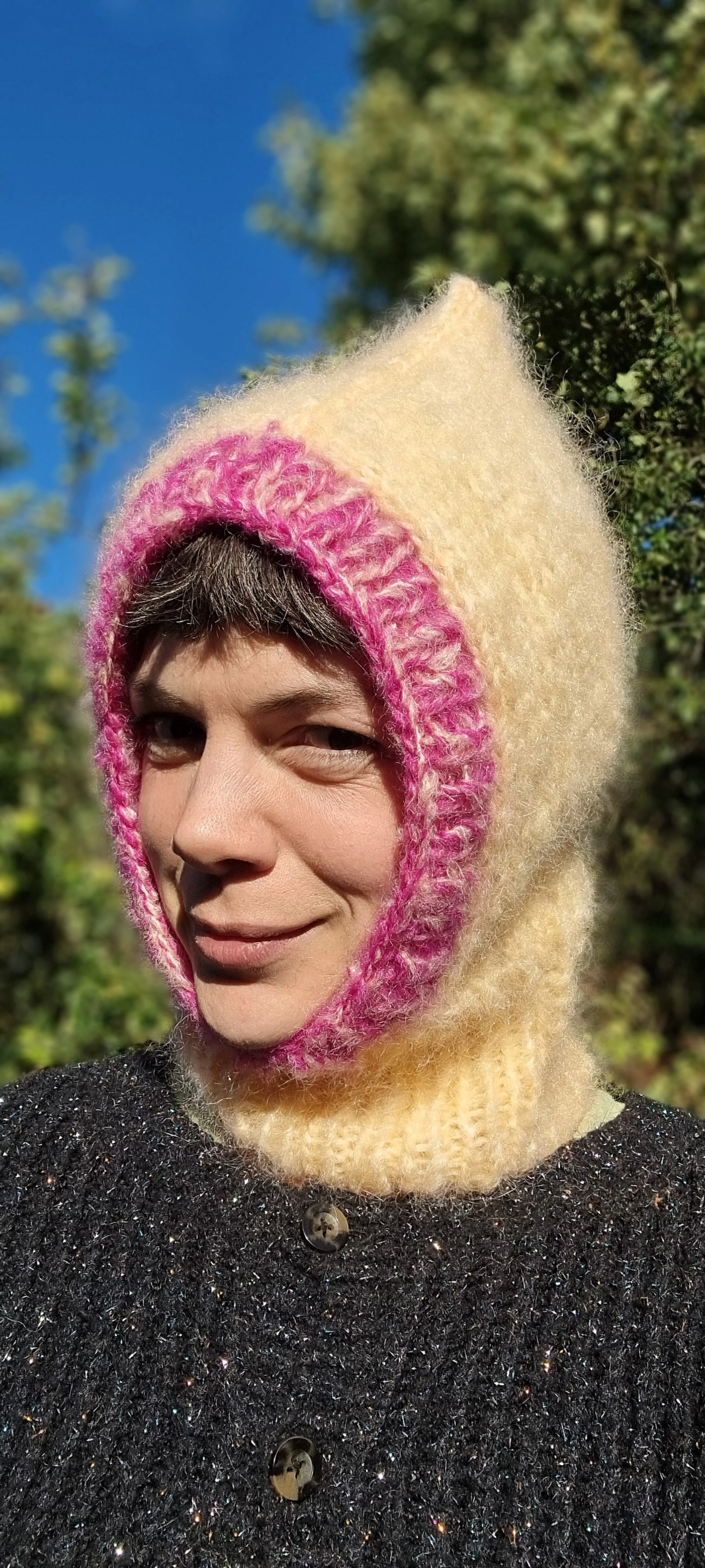 Person outdoors wearing a peach and pink knitted balaclava with a pixie point, smiling slightly, with blurred green trees in the background and a blue sky.