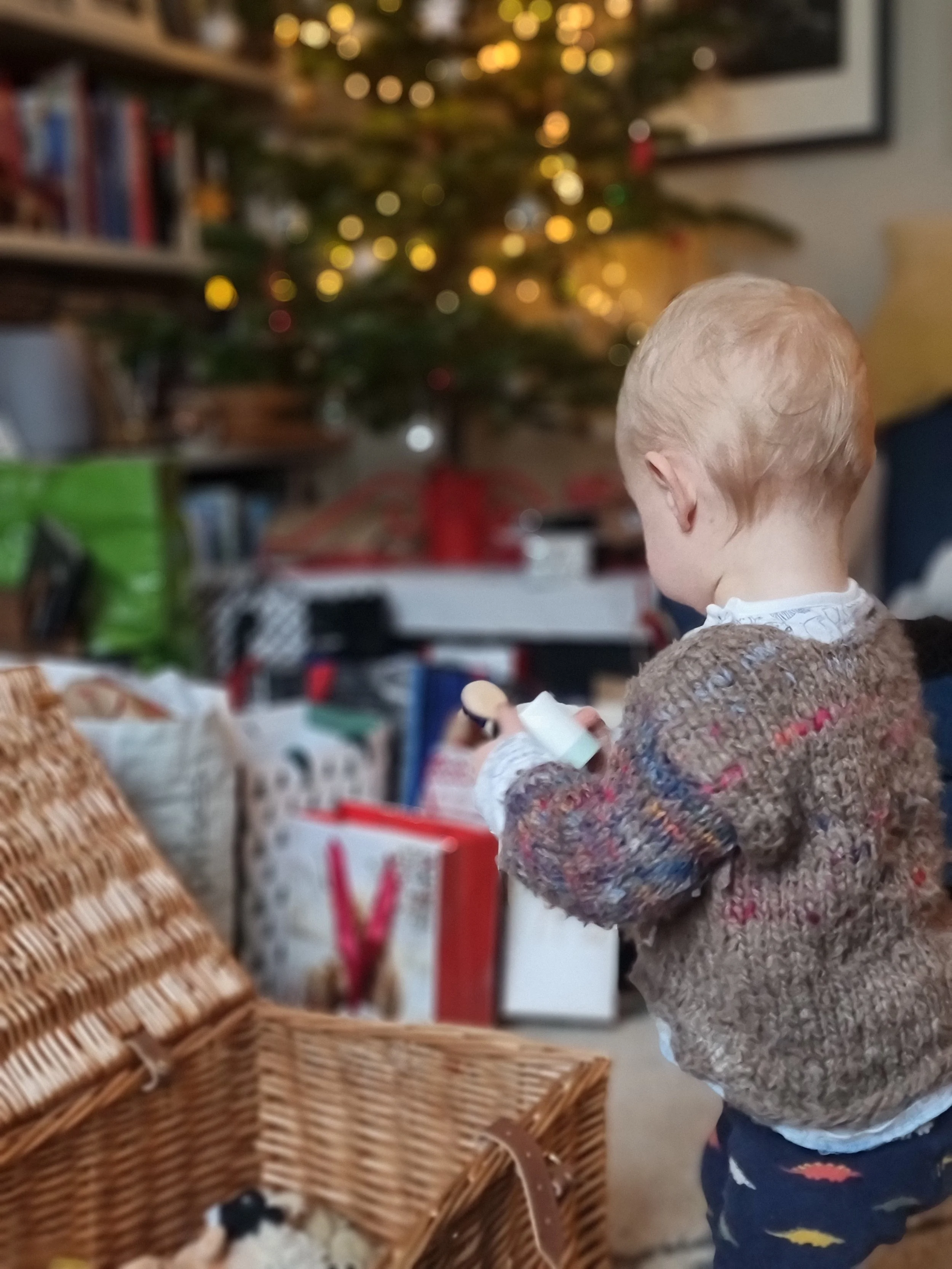 Young child with blonde hair wearing a colorful sweater, holding a toy or object, standing in a room decorated for Christmas with a decorated Christmas tree and wrapped presents in the background.