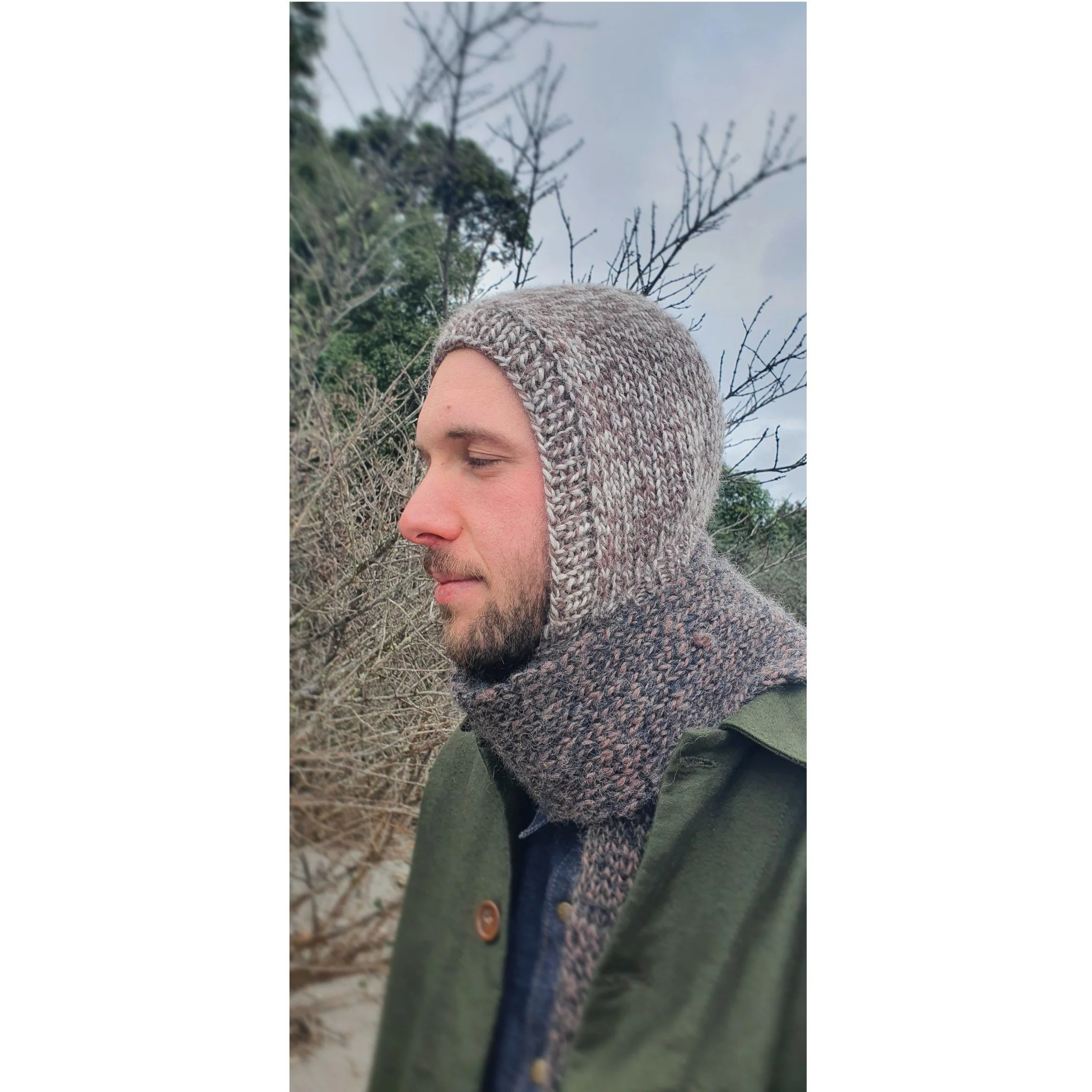 A man with a beard wearing a knit winter balaclava and a matching scarf outdoors, with leafless trees in the background.