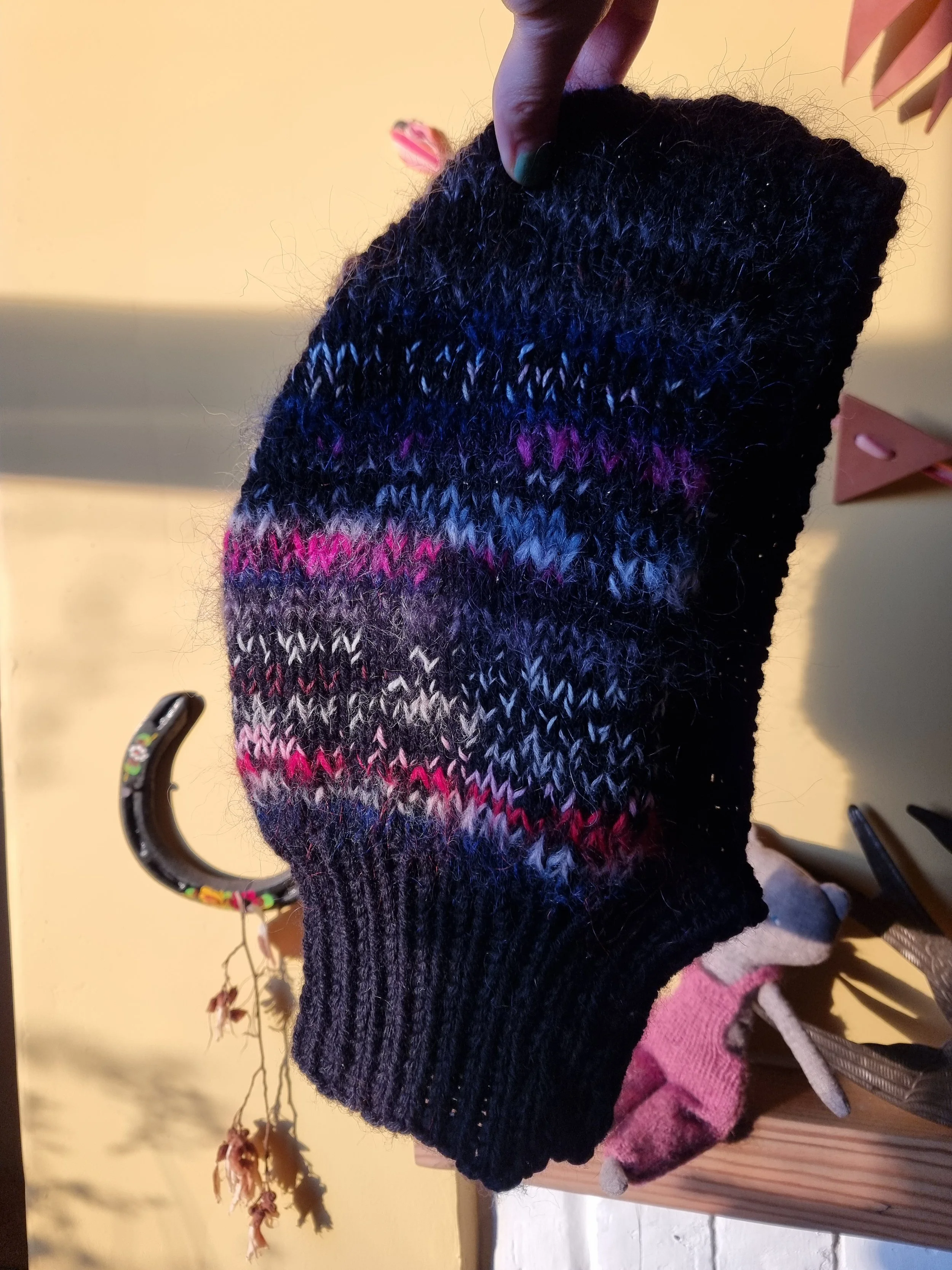 A knitted black balaclava with colorful stripe patterns in pink, purple, and gray, hanging on a hook.