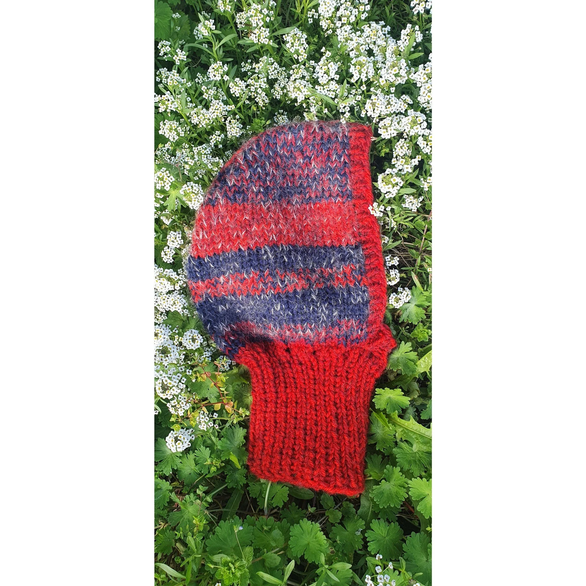 A multicolored knit mitten with red, navy, and gray stripes resting on a bed of small white flowers and green foliage.
