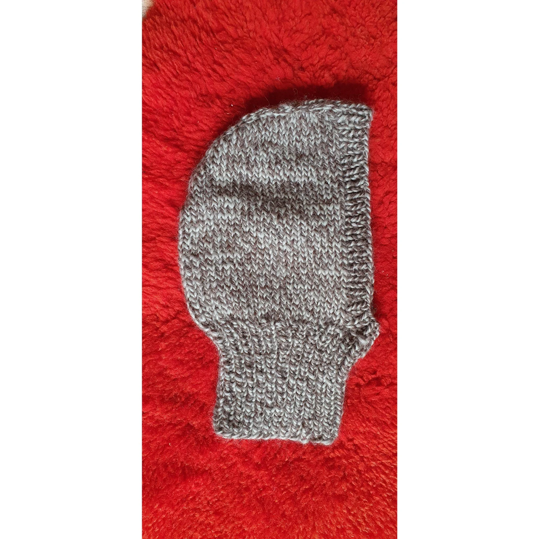 Gray knitted balaclava with white and black flecks, resting on a red fuzzy surface.