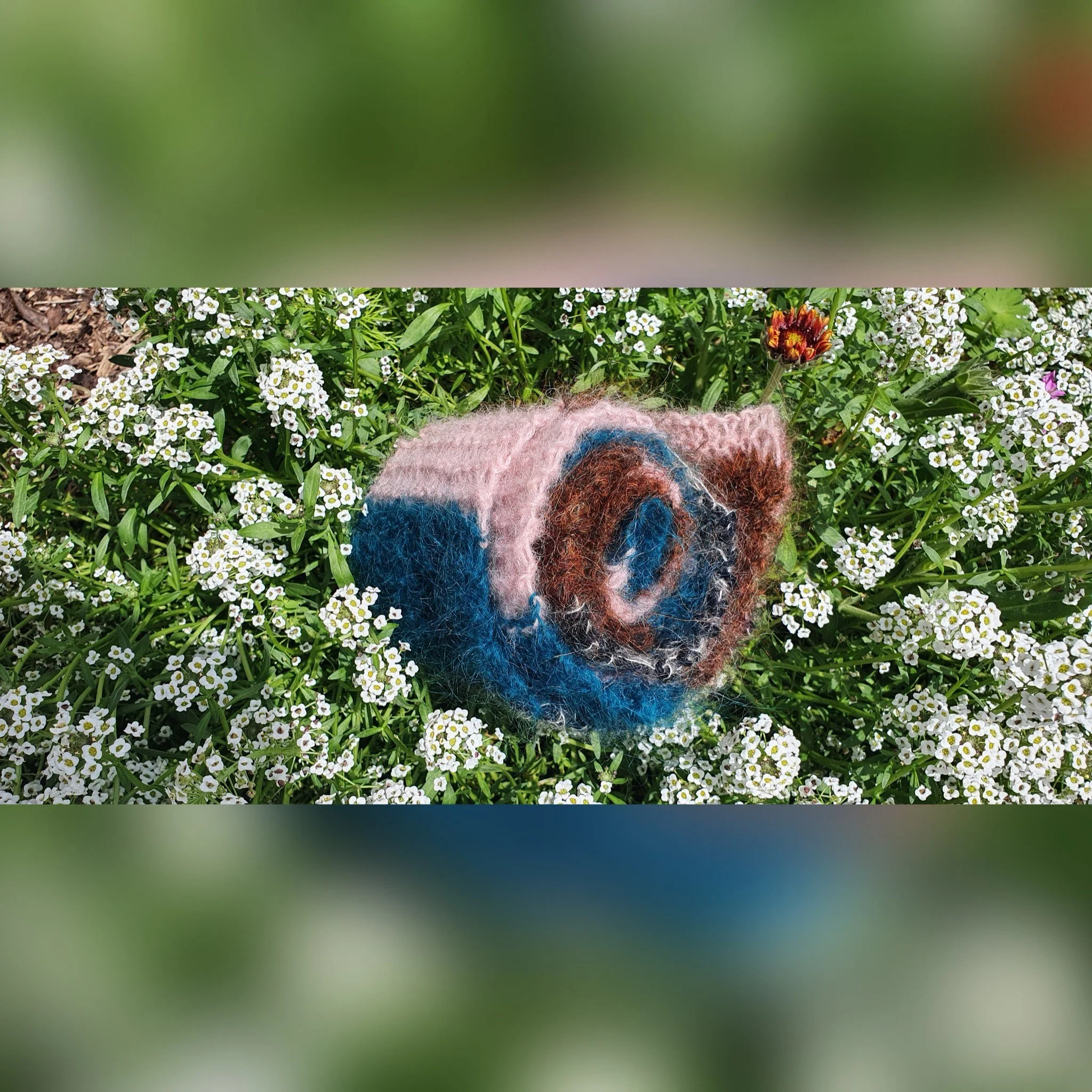A colorful, fluffy, felted wool scarf with pink, blue, brown, and black colors resting among white flowers in a garden.