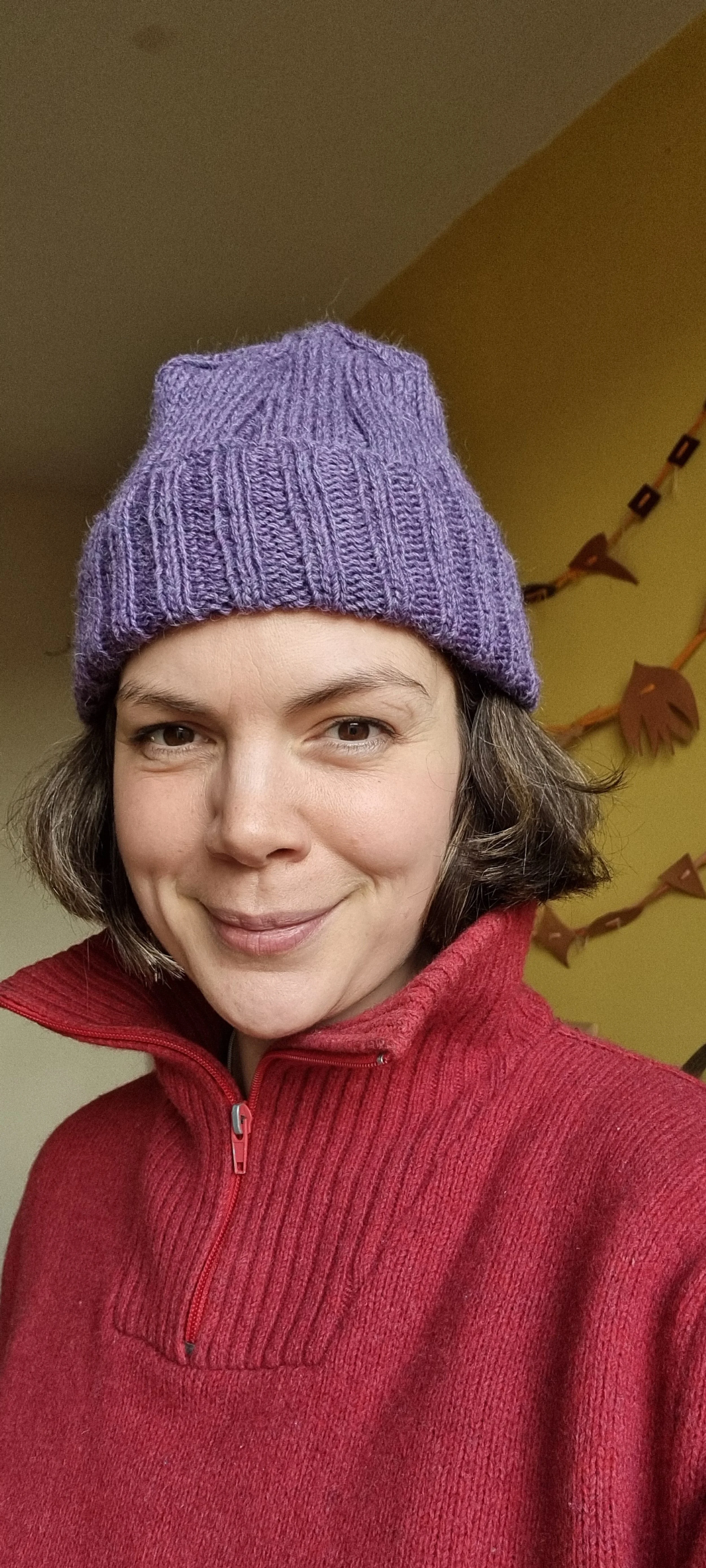 A woman smiling wearing a purple knit hat and a red zip-up sweater, with colorful paper leaf decorations in the background.